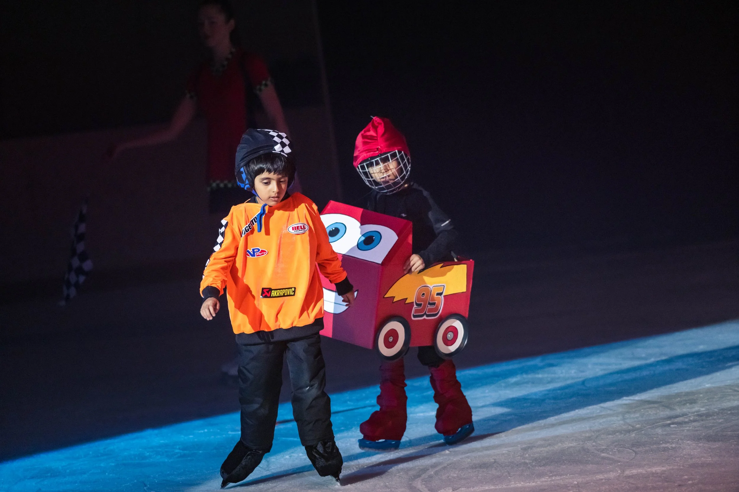 Two children ice skating at night, one dressed as Lightning McQueen from Cars and the other in an orange jacket and black pants, pulling a cart shaped like a cartoon character with big eyes and the number 95.