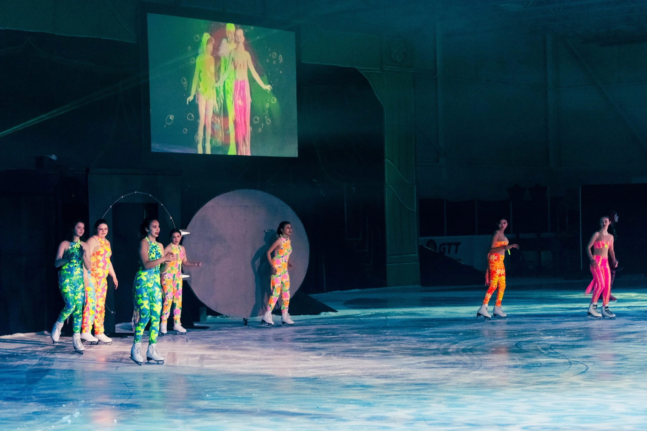 A group of young women figure skaters dressed in colorful tropical-themed costumes are performing on an ice rink. There is a large screen projecting their image above them and a dark background surrounding the scene.