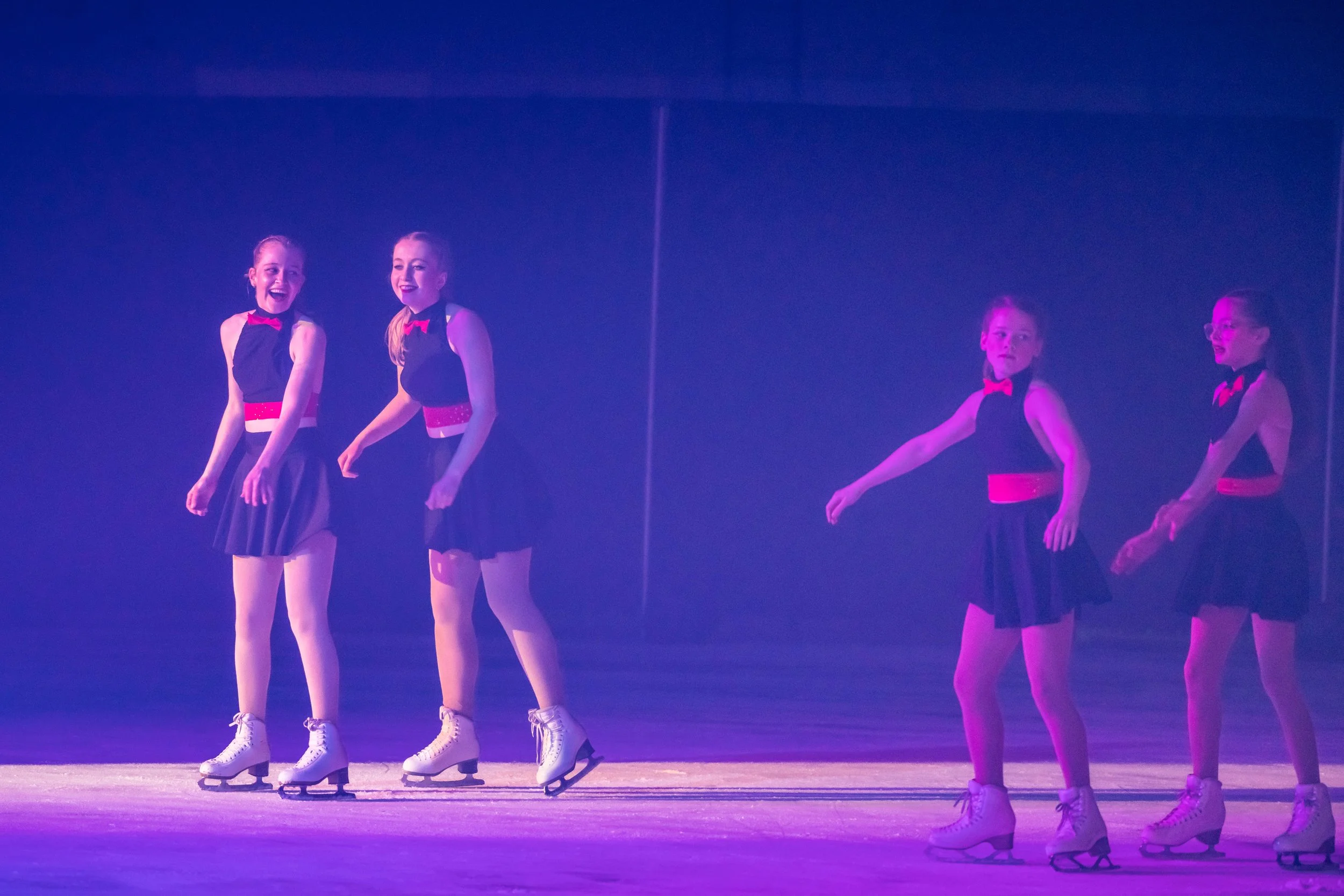 Four young girls wearing black with red accents figure skating on ice under dim purple lighting.