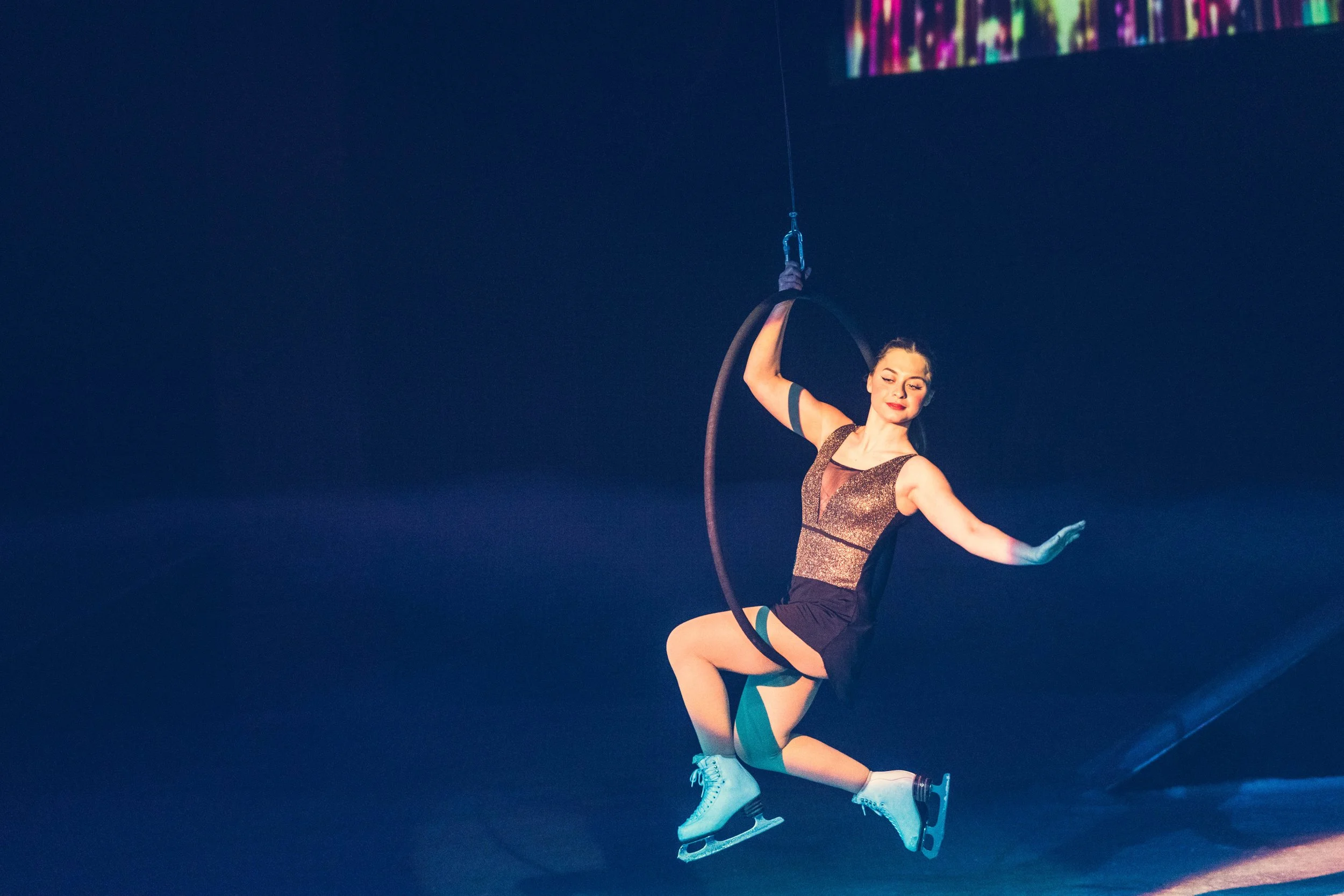 A female acrobat in a glittery costume performing on an aerial hoop, wearing ice skates, against a dark background with colorful projections.