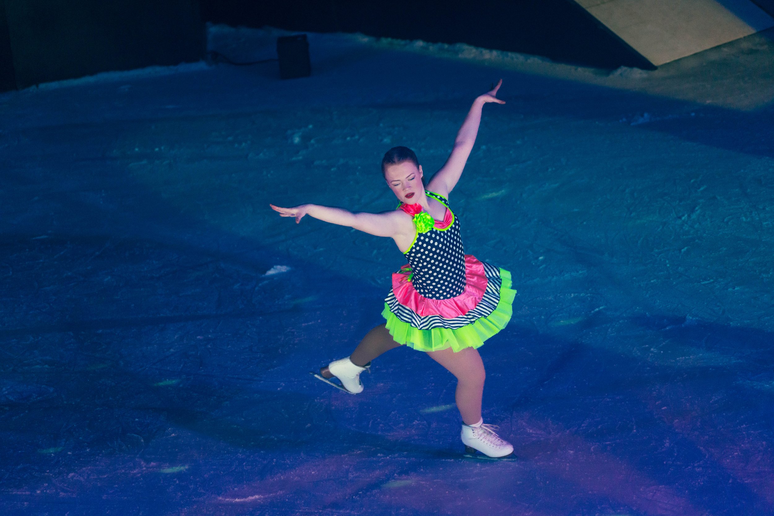 A female figure skater performing on ice, wearing a colorful costume with a polka dot top, neon green and pink accents, and a frilly skirt, with her arms raised and eyes closed.