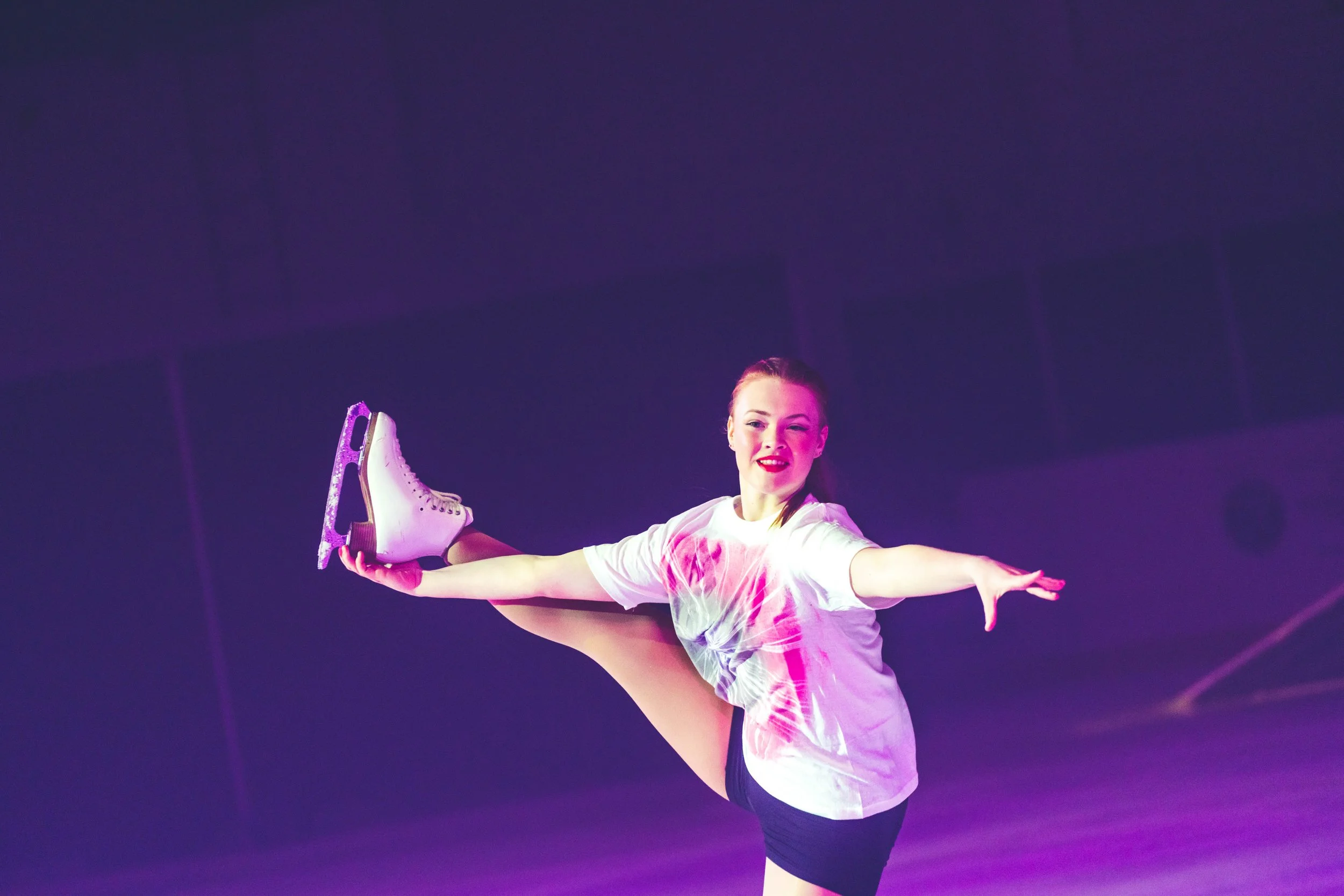 A young woman in a white T-shirt and black shorts performs a spiral dance move while holding white and purple ice skates, with a dark purple stage background and stage lighting.