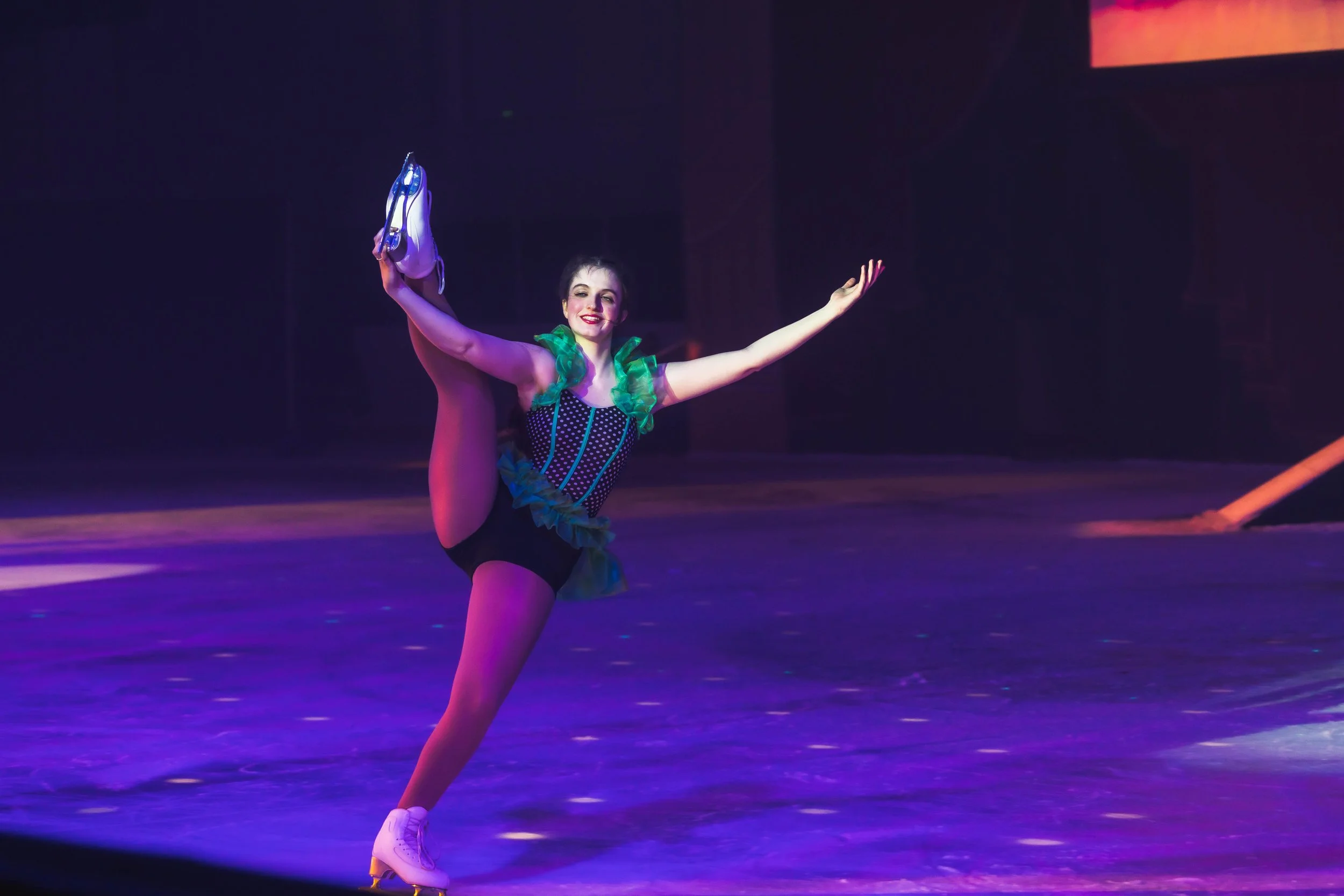 A female figure skater performing a move on the ice, holding a skate in one hand and extending the other arm outward, dressed in a colorful costume with green and purple accents.