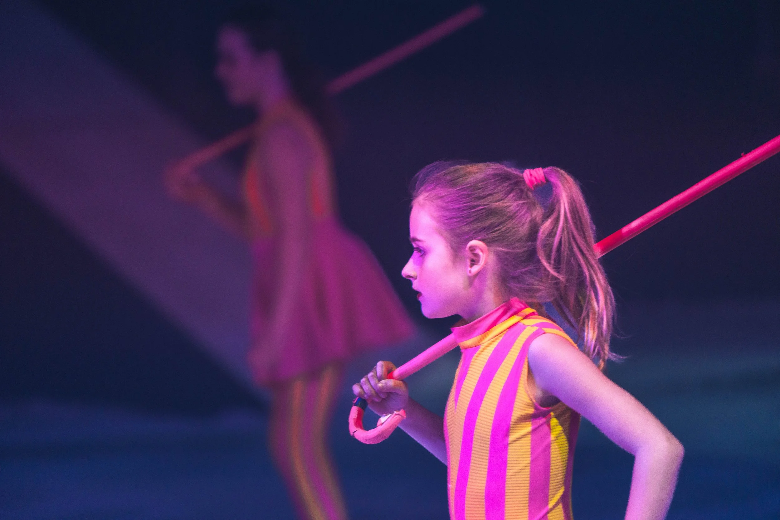 A young female gymnast in a pink and yellow striped uniform holding a pink pole, with a queen-like figure in a pink dress in the background under purple lighting.