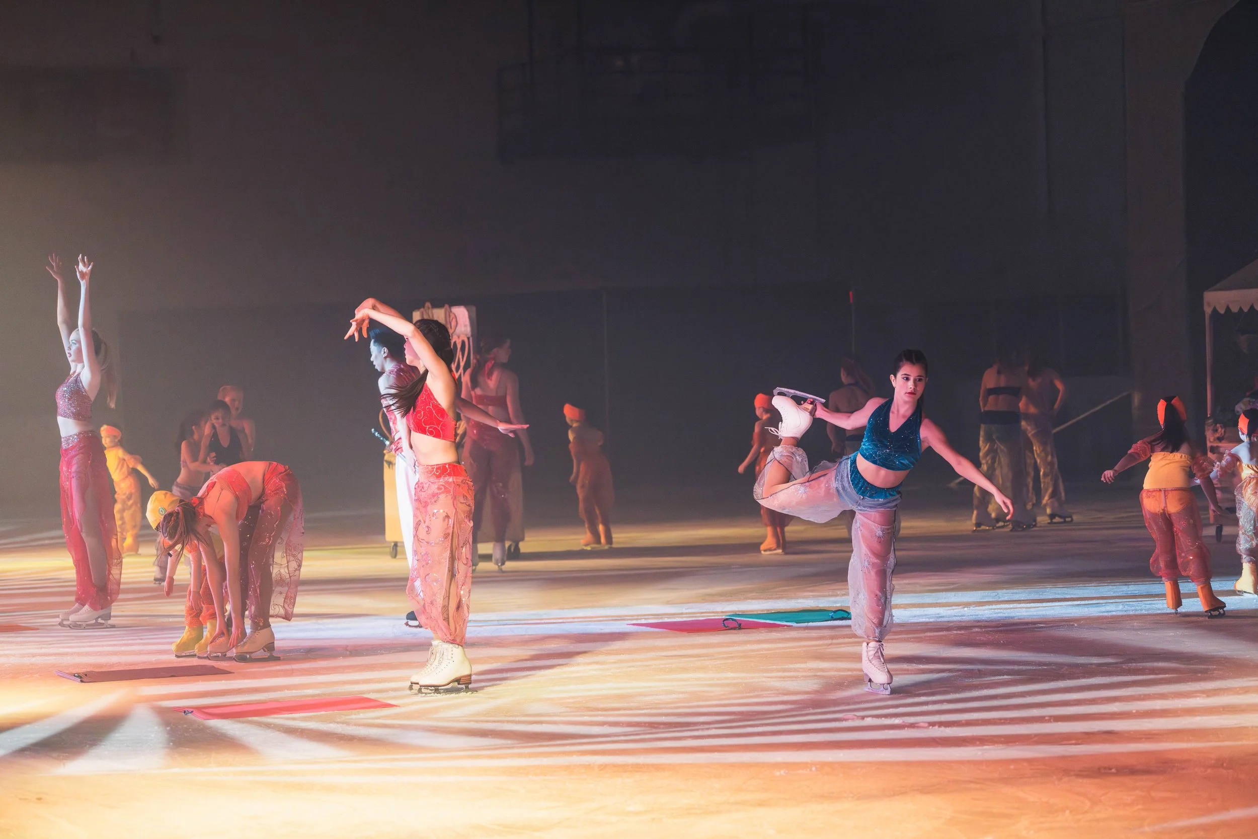 Group of ice skaters practicing on an indoor ice rink, some are standing, others are performing spins or bends, with a dark background and warm lighting.