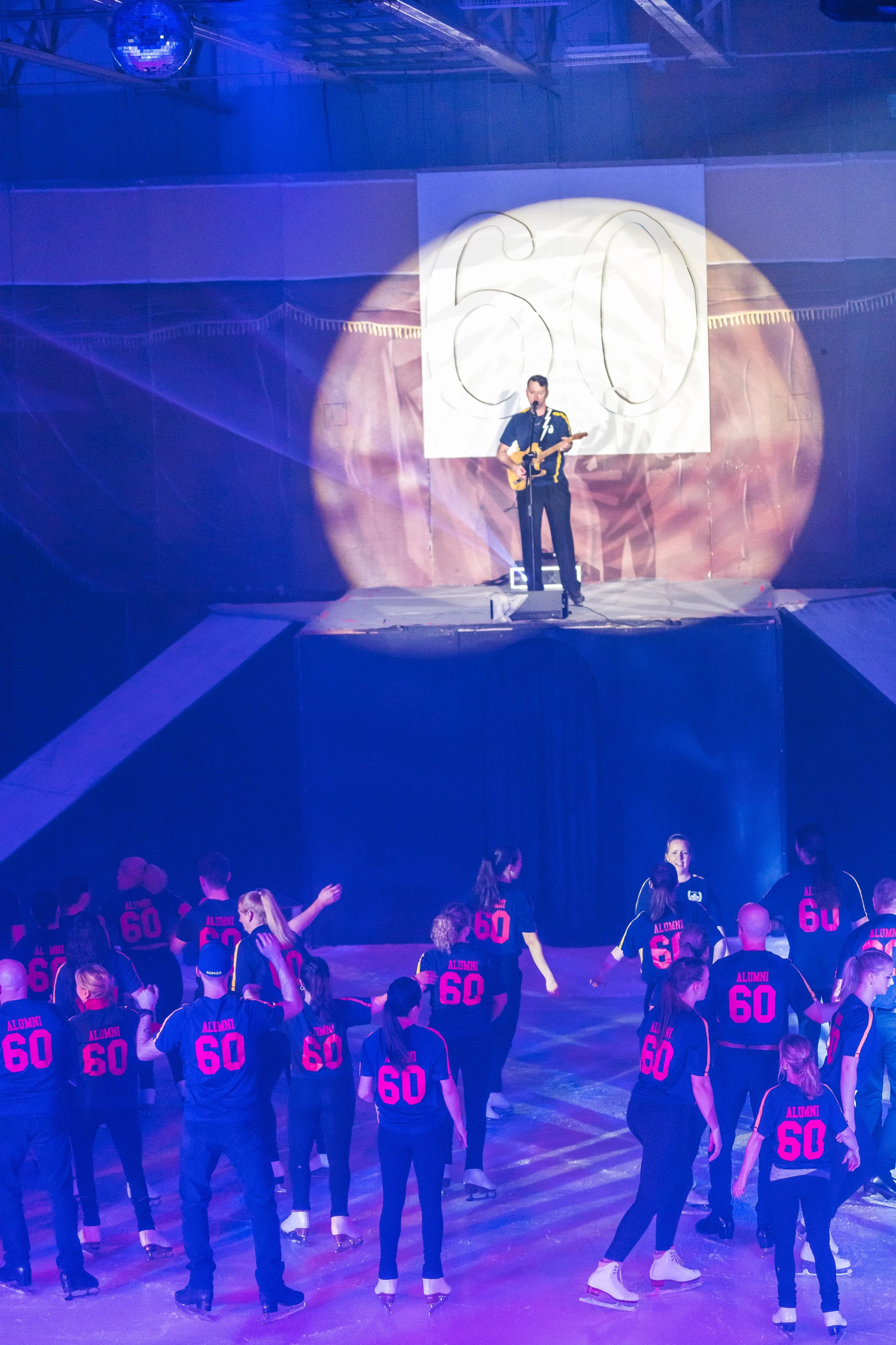 People on ice skating rink wearing matching black shirts with pink 'ALUMNI 60' text, watching a performer playing guitar on stage with a large '60' displayed behind him.