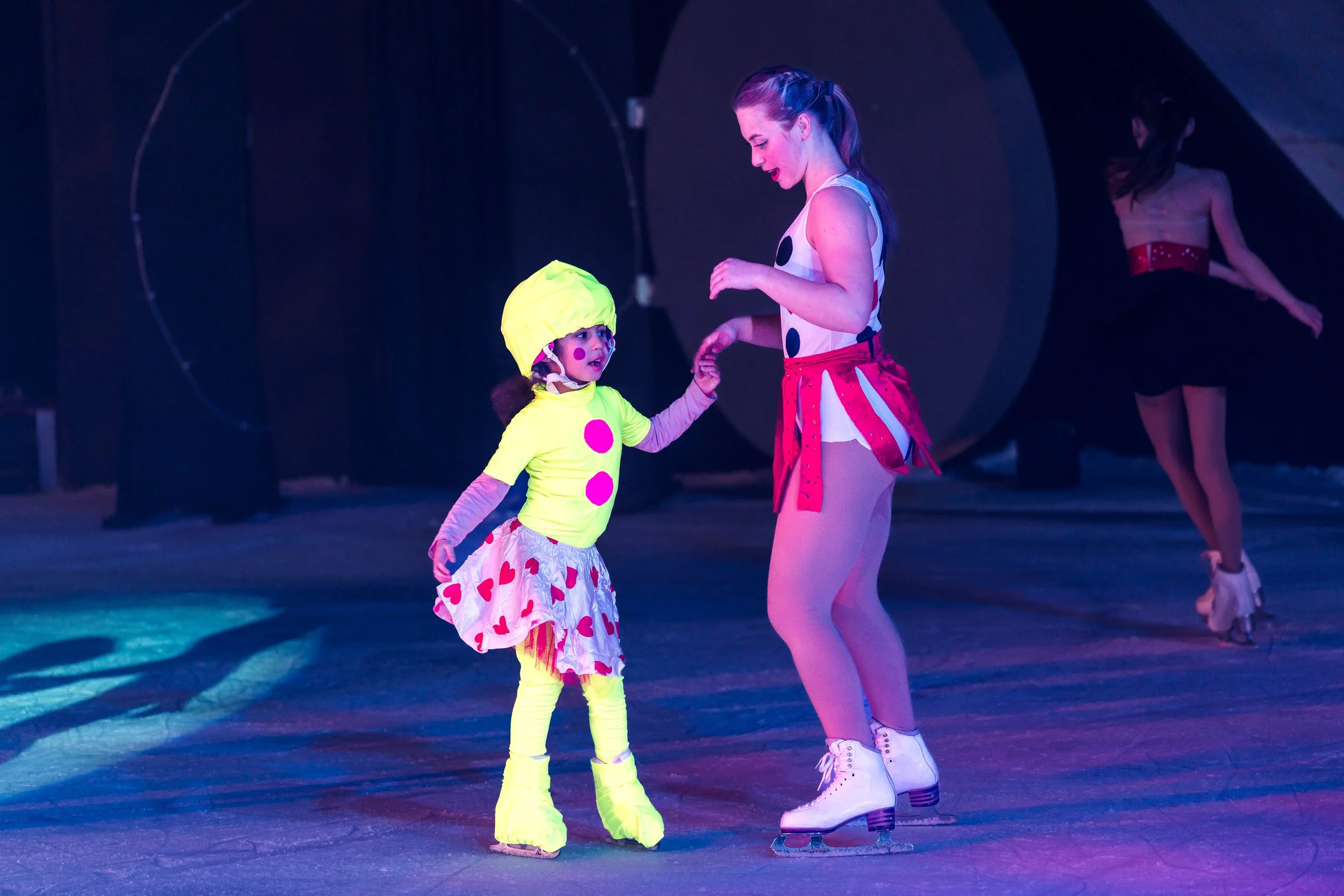 A young girl dressed in a colorful clown costume with yellow tights, a pink polka-dotted skirt, and a bright yellow hat, holding hands with a woman in a polka dot and red skirt costume, both wearing ice skates on an ice rink, during a performance.