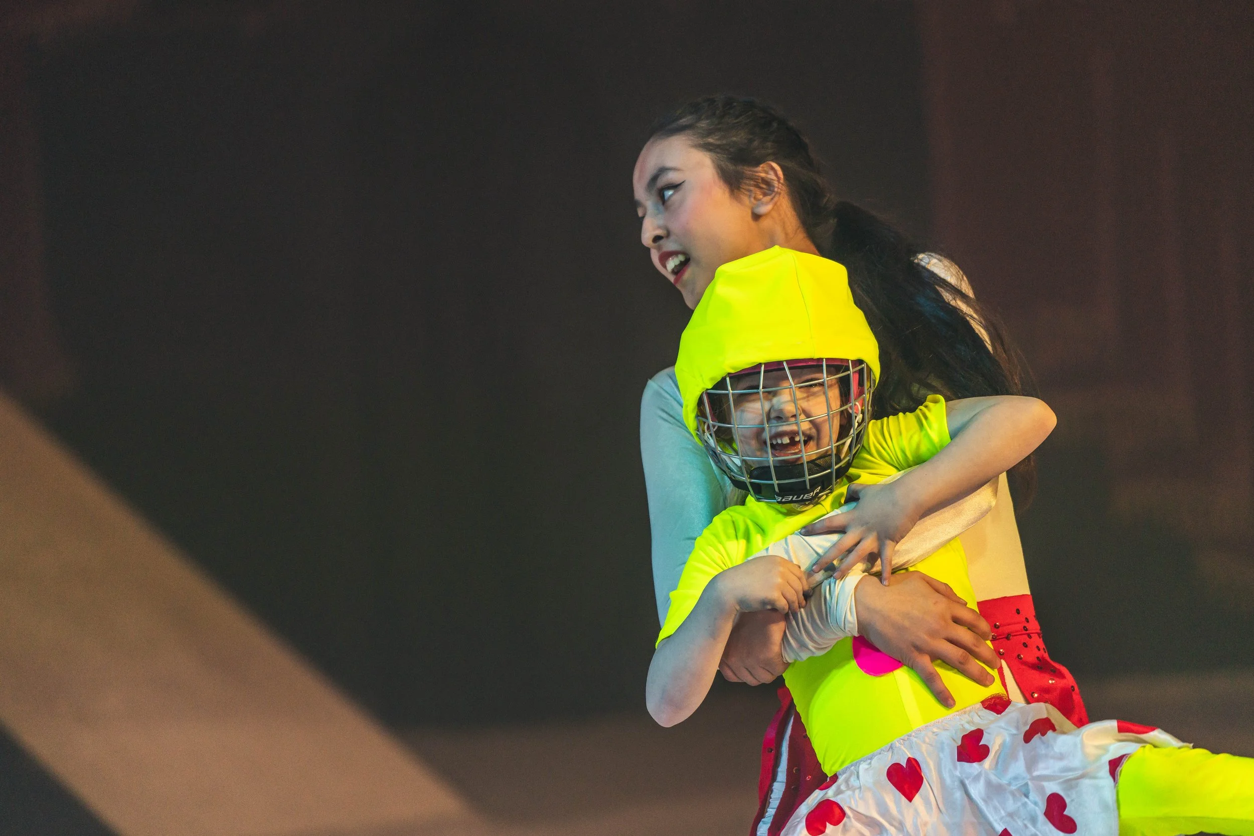 A woman dressed in bright sportswear is embracing a young girl dressed as a hockey goalie, both smiling. The girl is wearing a yellow goalie mask and jersey, and the woman has her hair tied back.