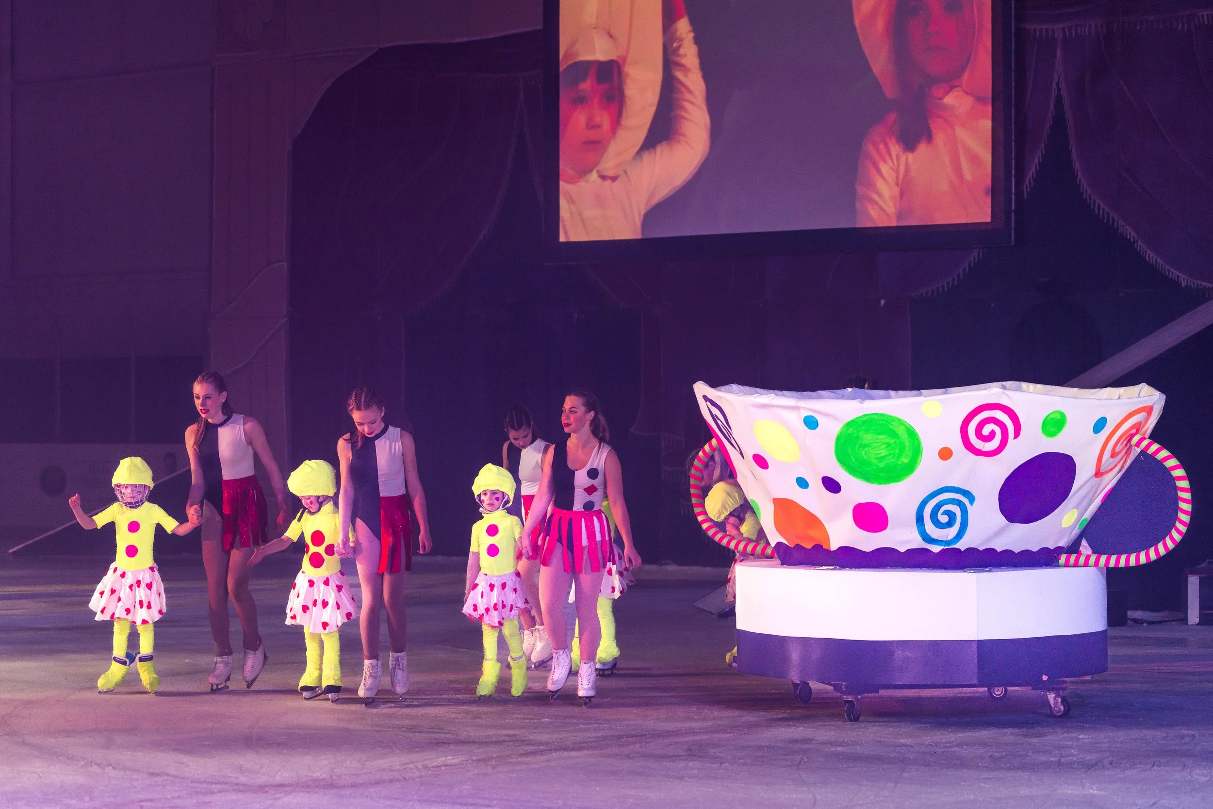 Children dressed in colorful costumes with polka dots and bright yellow hats, participating in a stage performance or dance routine on a dark stage, with a large, multicolored, spiral-patterned prop also on stage.