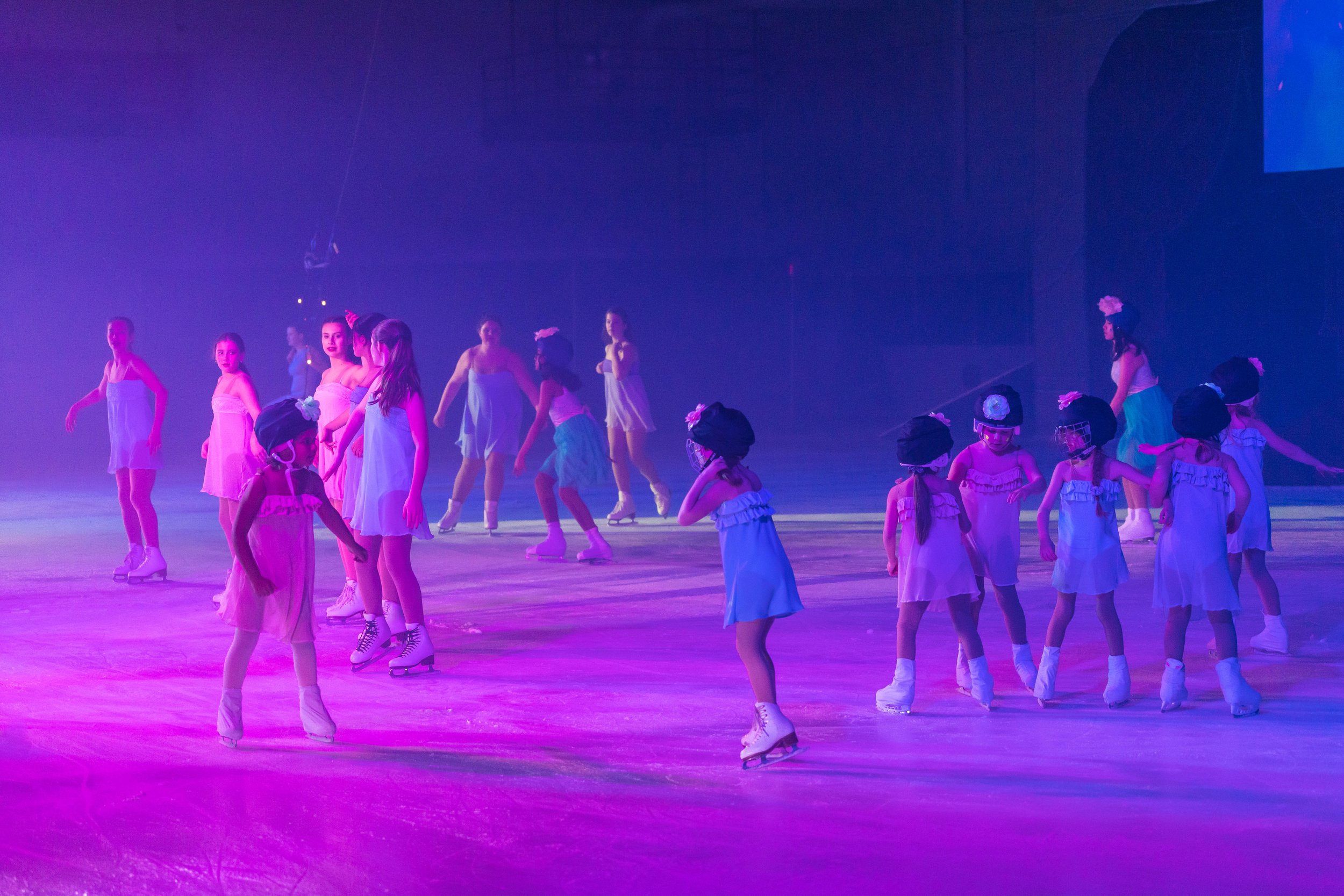Children ice skating on an indoor rink with purple and pink lighting, wearing pastel-colored dresses and ice skates.