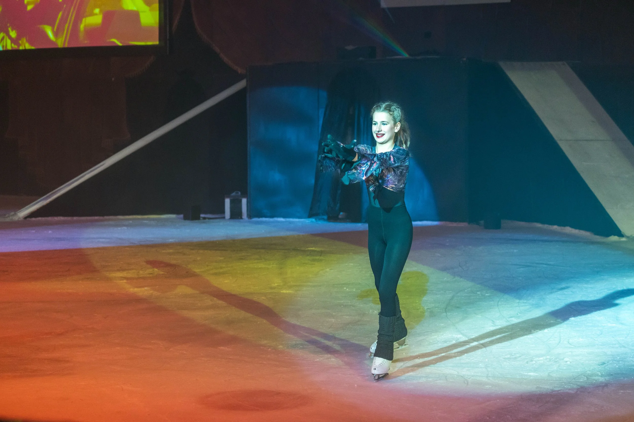 A young woman roller skates on an indoor rink, smiling and wearing a colorful, shiny jacket.