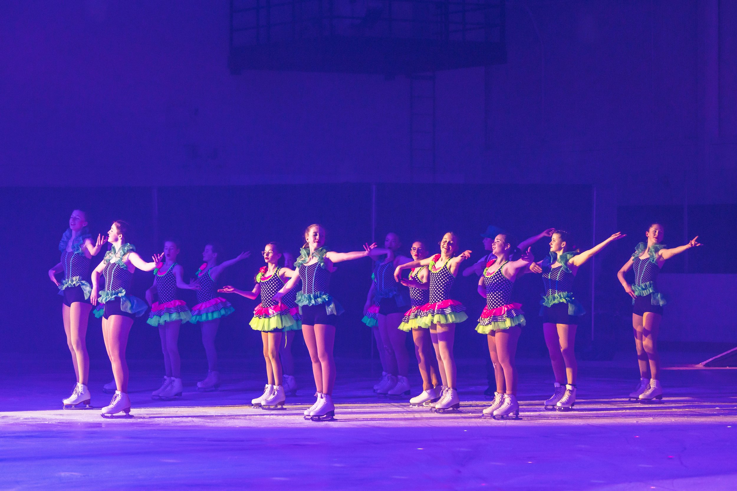A group of female figure skaters performing a synchronized routine on ice, wearing colorful costumes with polka dots and ruffled skirts, under purple stage lighting.