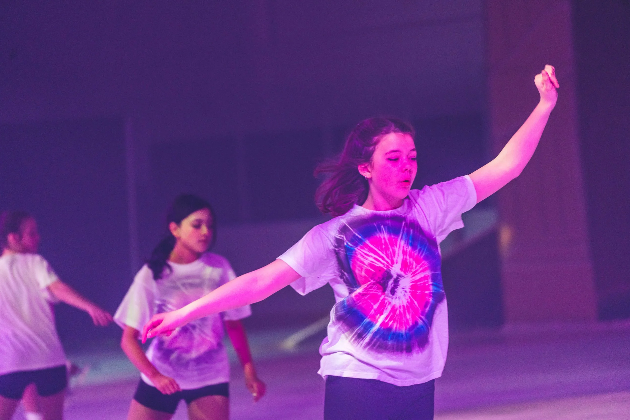 Women dancing in a group with colorful lighting, one woman in the foreground has arms outstretched and is wearing a tie-dye t-shirt.