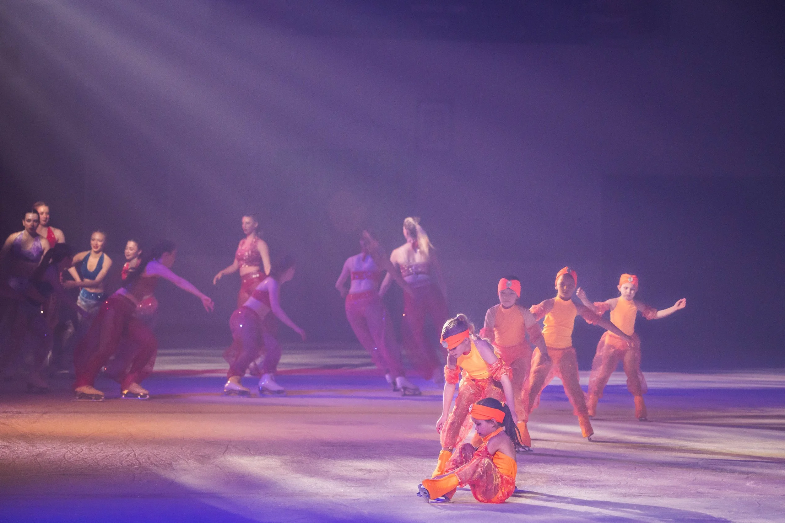 A group of dancers on stage, with some wearing colorful costumes, performing under purple stage lighting.