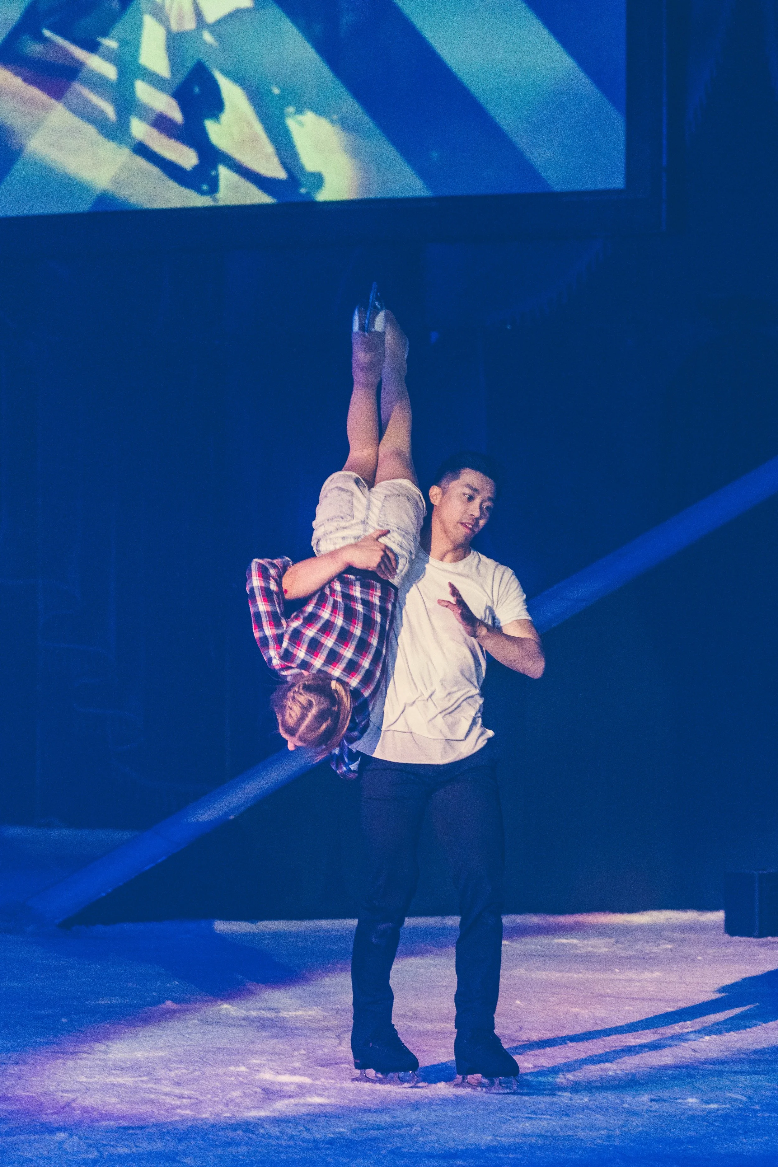 A person wearing ice skates holding another person upside down on the ice rink. The person on top is turned upside down, while the other supports them, wearing a plaid shirt. The scene is illuminated with blue stage lighting and shows a reflective su