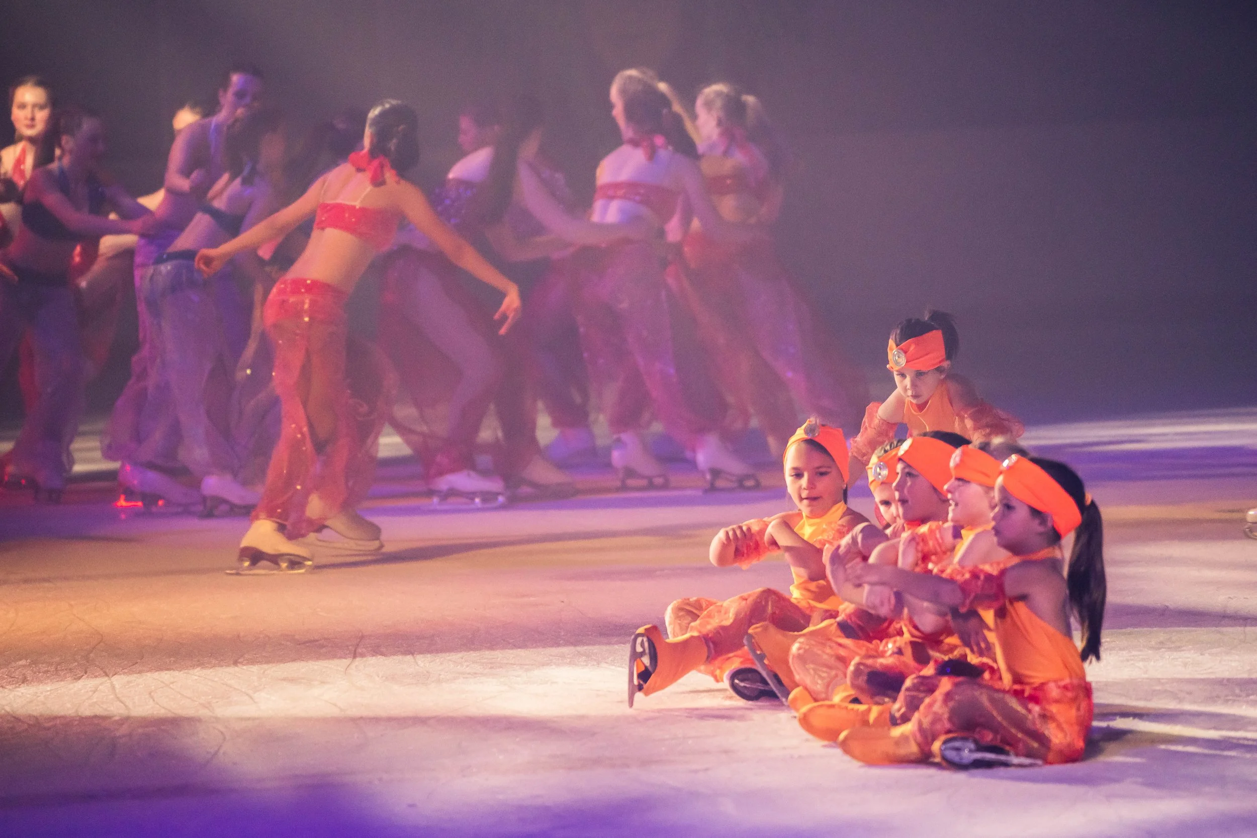 Children on roller skates dressed in orange costumes sitting on the ice rink with some children standing and talking in the background.