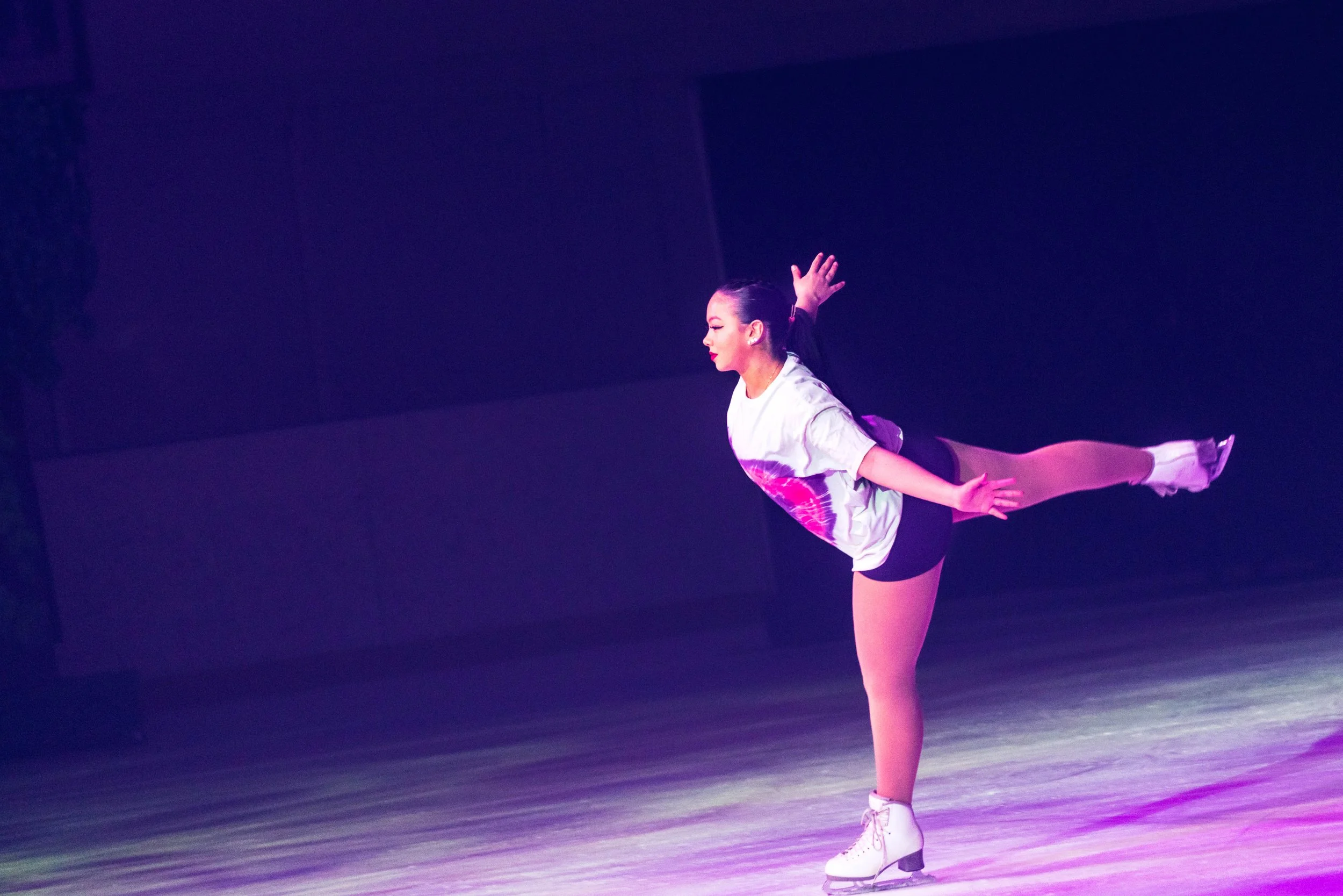 A female figure skater in a white and pink costume practicing on the ice rink with one leg extended behind her in a spiral position.
