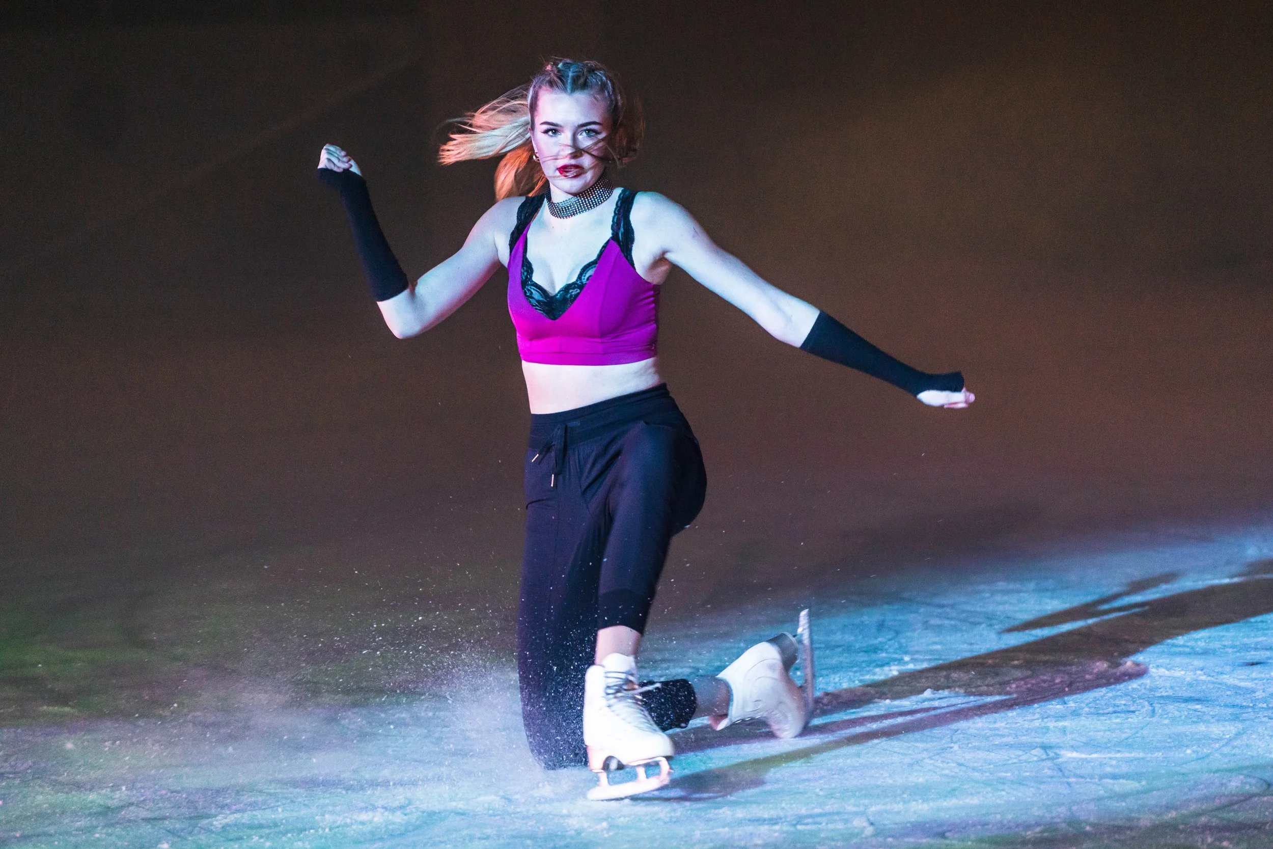 Young woman with blonde hair rollerblading at night on a dark street, wearing a pink top, black pants, and long black gloves, with a determined expression.
