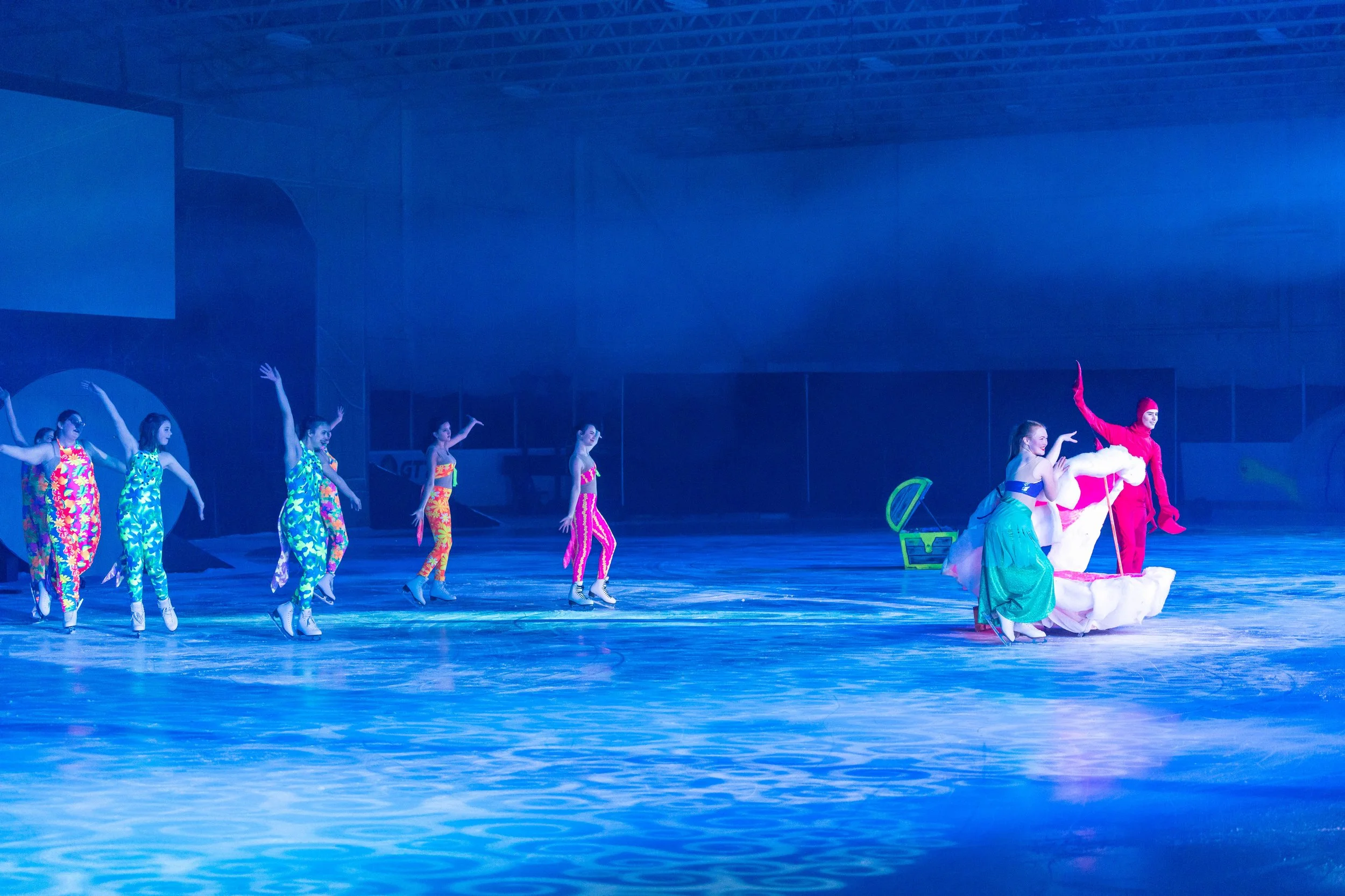 Ice skaters performing a synchronized routine on an ice rink with blue lighting, including a person dressed as a clown in a pink costume and a large white hat.