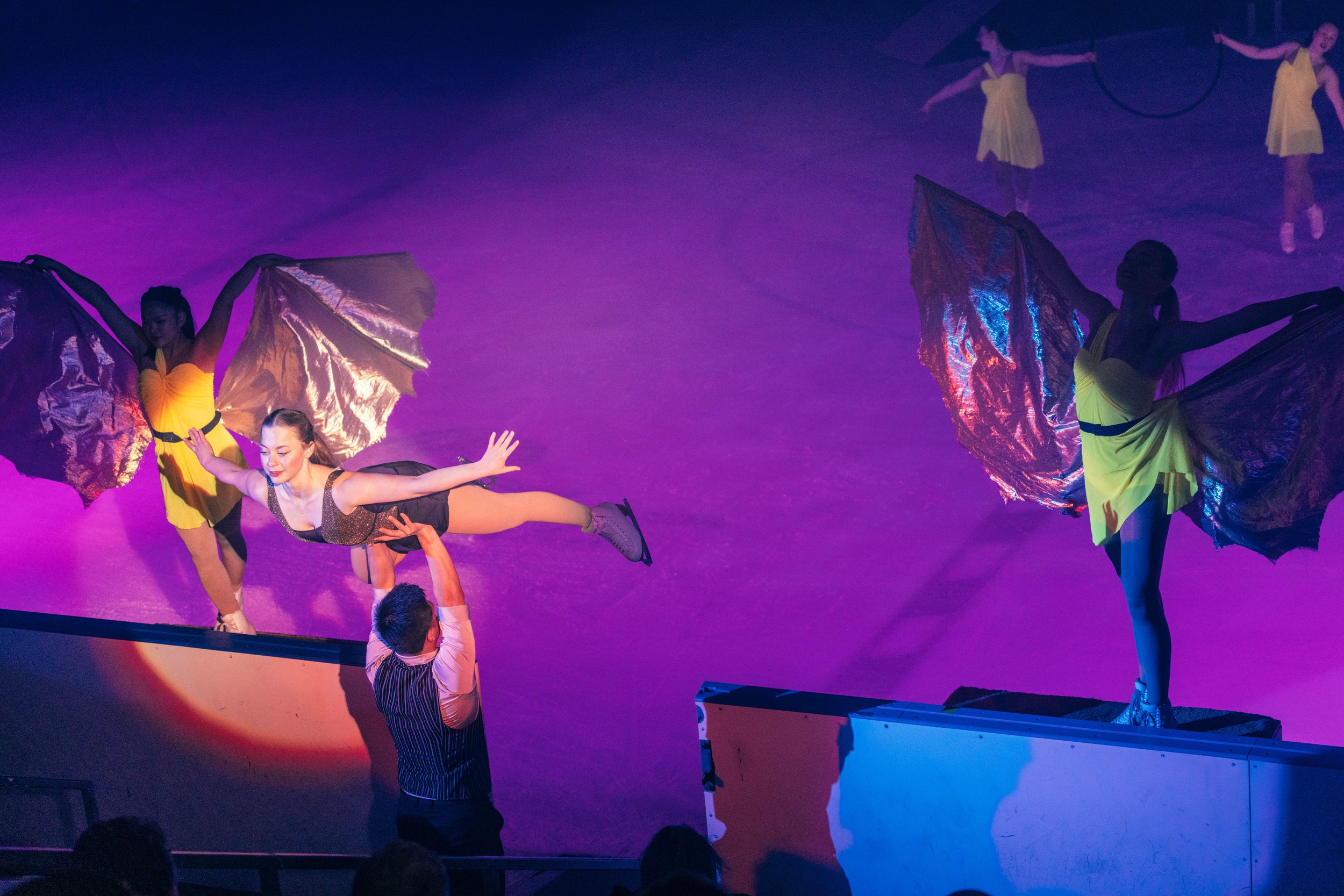 Circus performers, including acrobats, performing on stage with colorful lights and costumes, including a performer holding large metallic fabric wings.