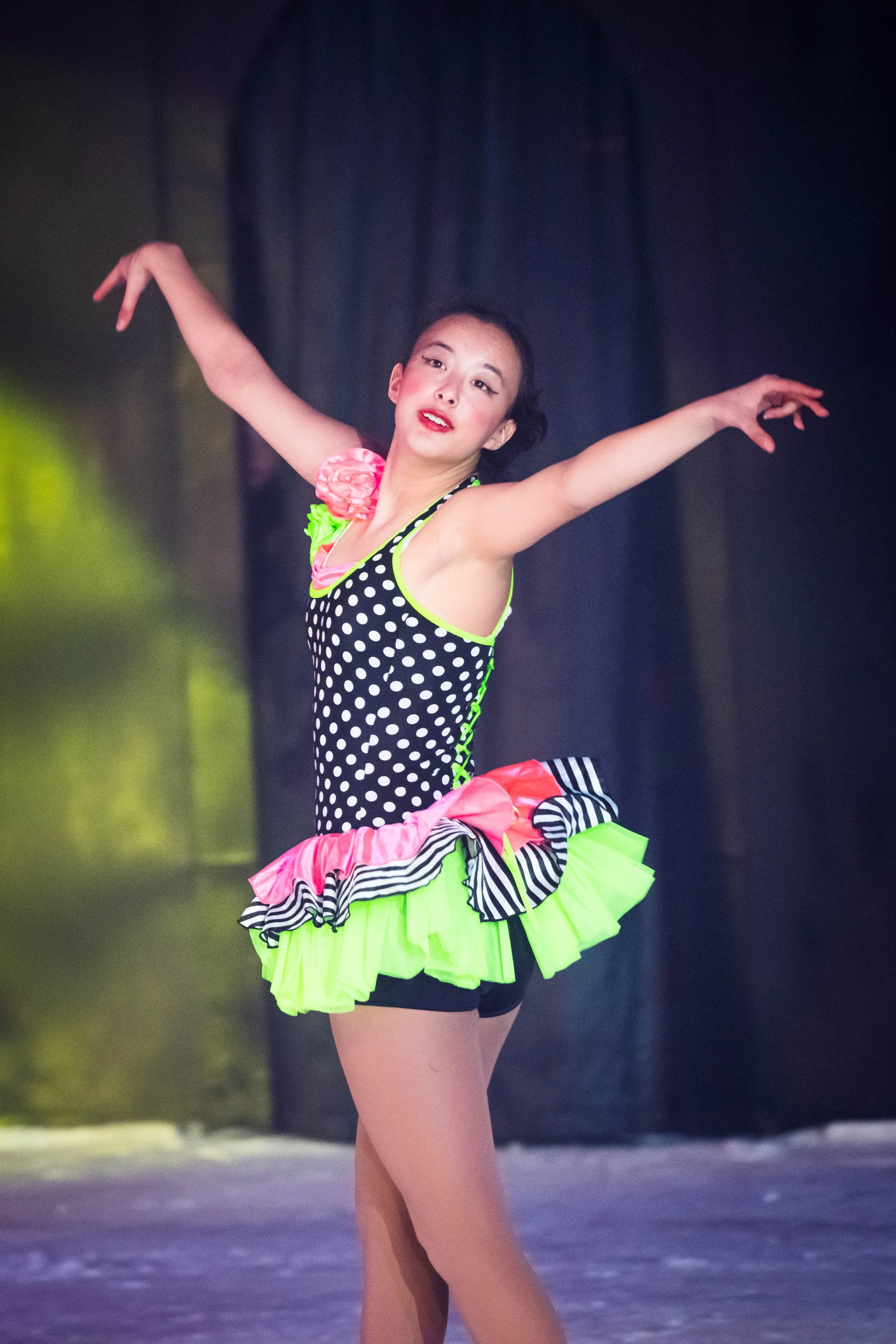 A young woman performing a dance on stage, wearing a colorful polka dot and neon green costume with frills and a large pink flower detail on the shoulder. Her arms are extended outward in a dance pose.