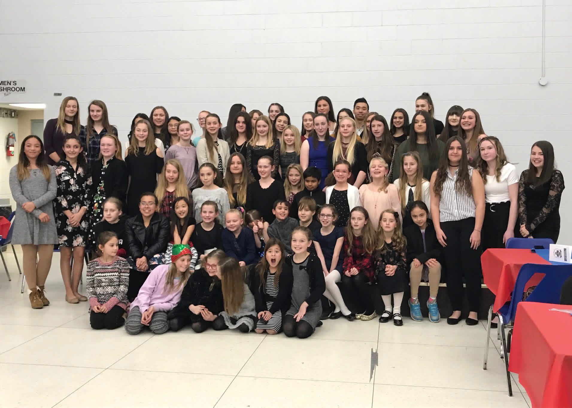 Group of children and teenagers posing for a photo at an indoor event, some sitting on the floor and others standing behind, with tables decorated for a celebration.