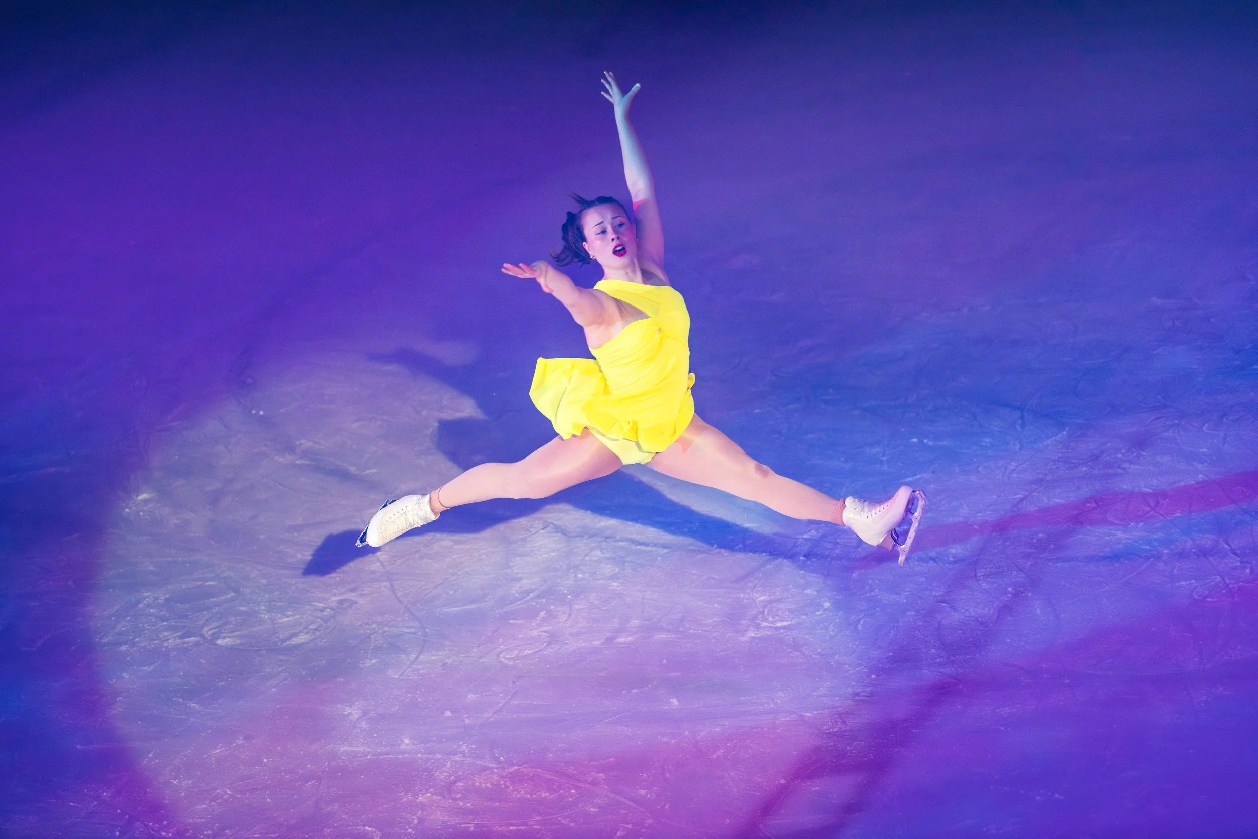 A female figure skater in a bright yellow dress performs an energetic move on the ice, with one leg extended and one arm raised above her head, in a colorful, illuminated ice rink.