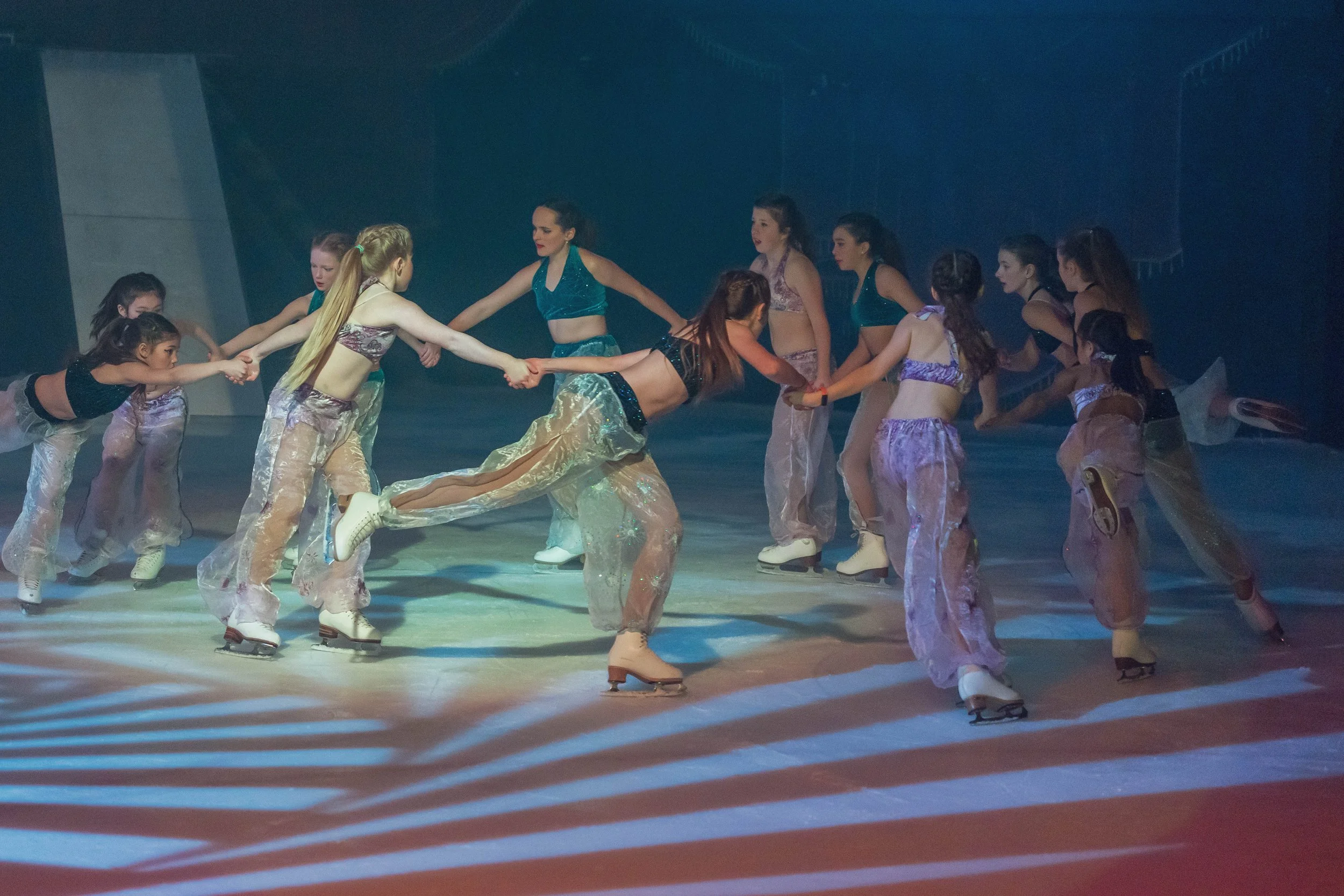 A group of young girls ice skating and holding hands in a circle during a performance or practice on an ice rink, with colorful lighting effects.