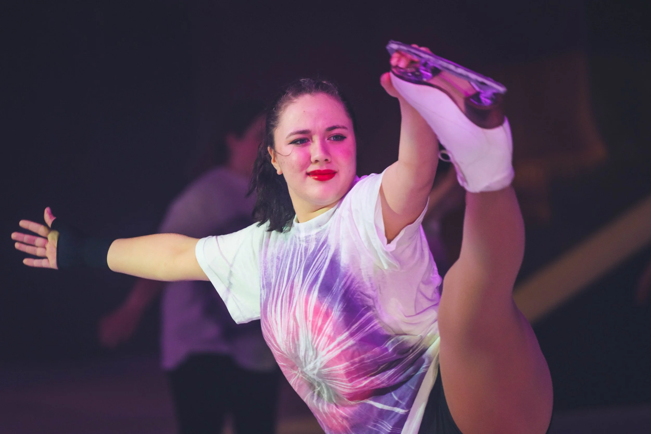 A young woman in a dance pose with one leg lifted and held by someone else, smiling, in a dimly lit environment with purple lighting.