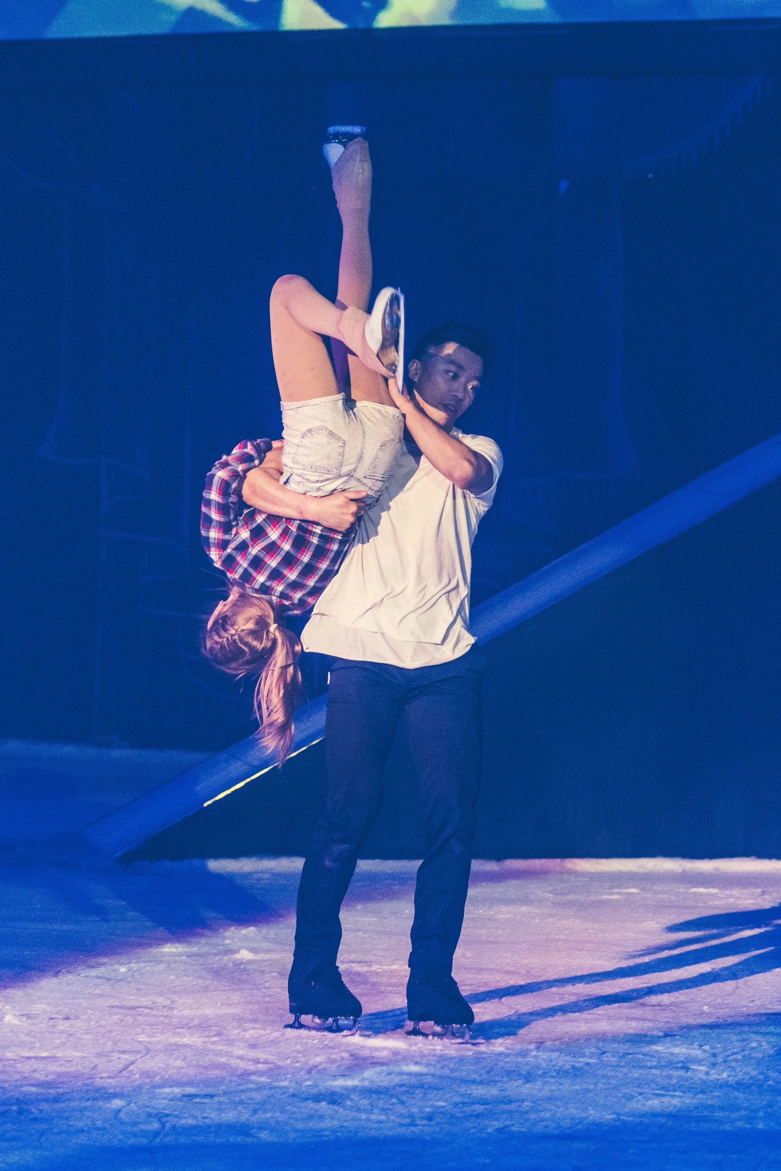 Person in ice skates being held by another person on an ice rink. The person being held is upside down, with legs in the air and head close to the ice. The background is dark with some structural elements visible.
