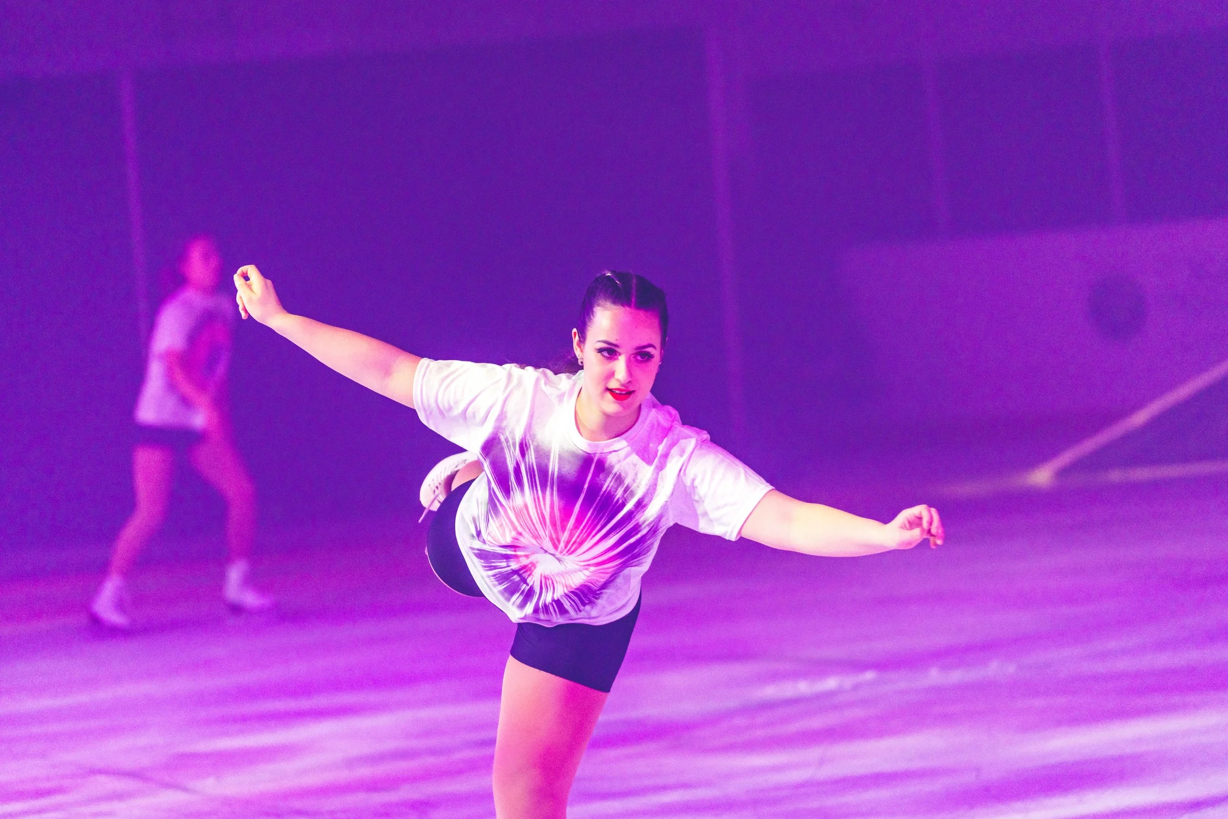 A young woman figure skater is performing a move on the ice rink, dressed in a white t-shirt and black shorts, with her arms extended and one leg lifted. She is focused and in motion, with a purple-lit background and another skater in the distance.