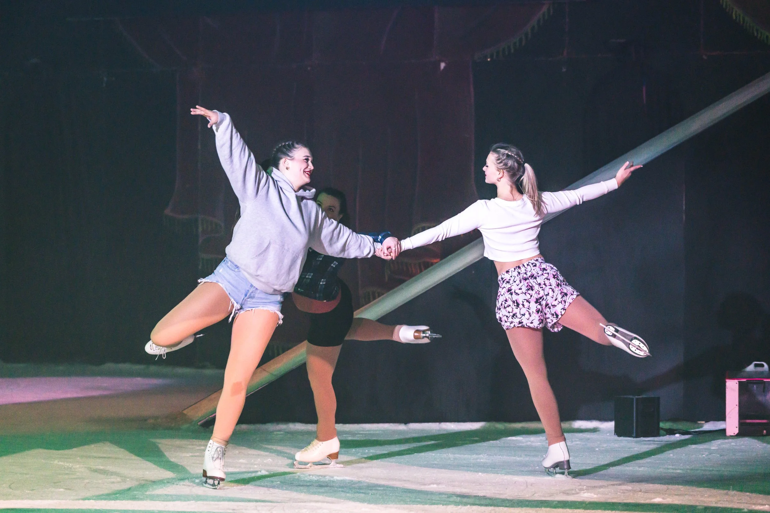 Two young women in figure skating outfits practicing on ice rink, holding hands and balancing on skates, with a coach or instructor in the background, colorful lighting creating a vibrant atmosphere.