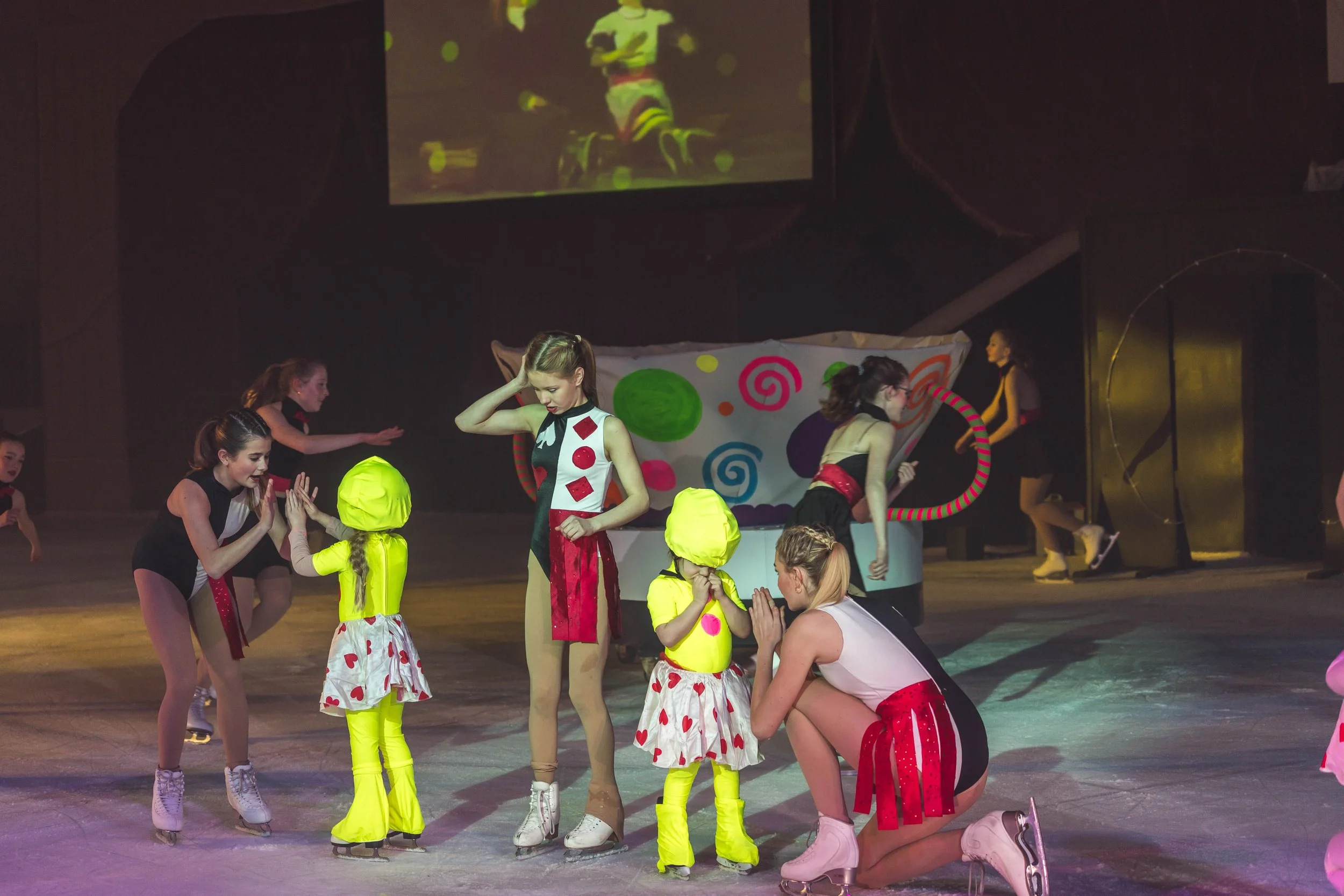 Children and young women dressed in colorful costumes and roller skates, performing in a circus or stage show with a colorful backdrop and a large screen projecting the performance.