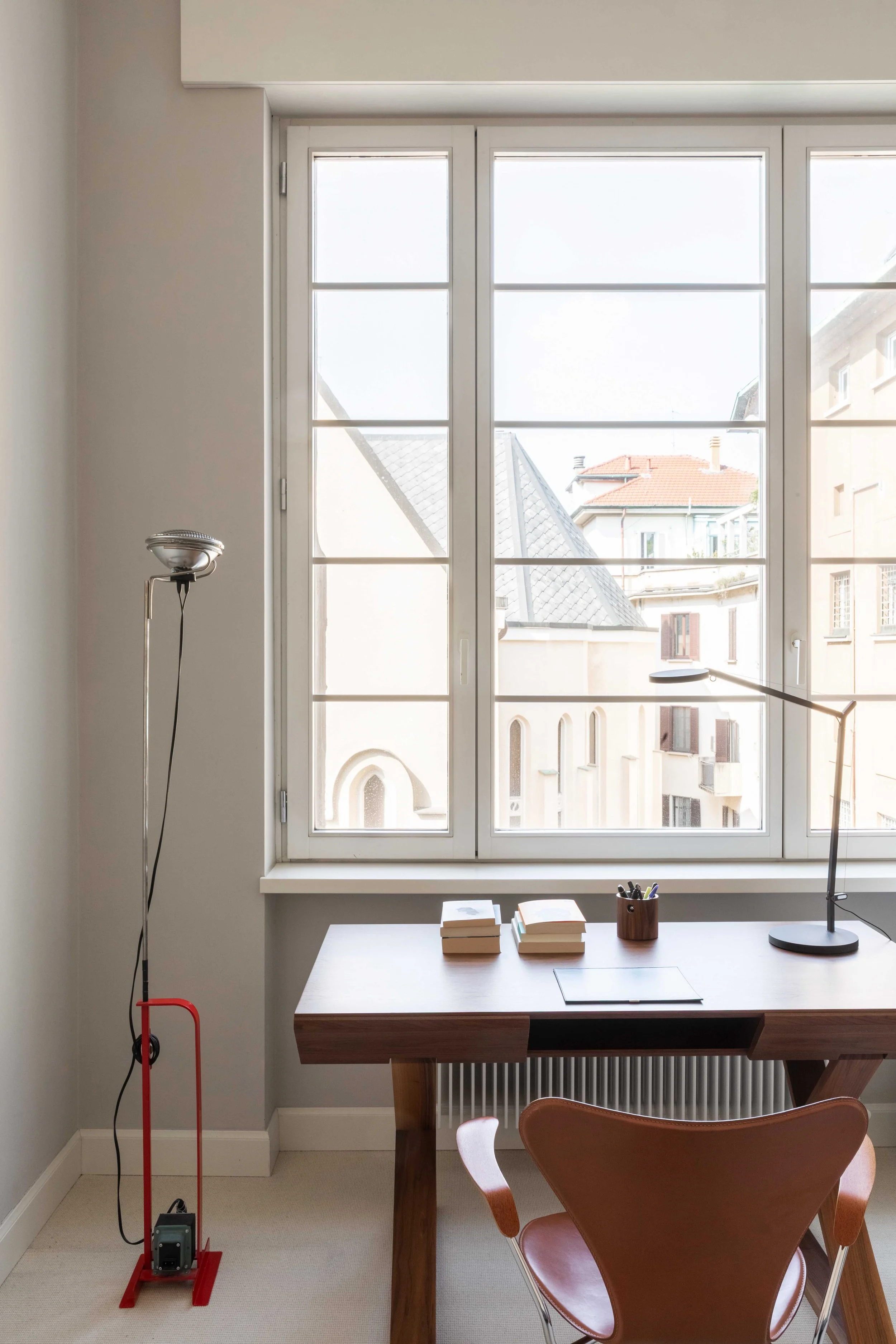 Ingresso di una stanza con una scrivania in legno, una sedia di pelle marrone, una lampada da tavolo, alcune riviste e penne, una finestra grande con vista su tetti e edifici, un termosifone sotto la finestra.