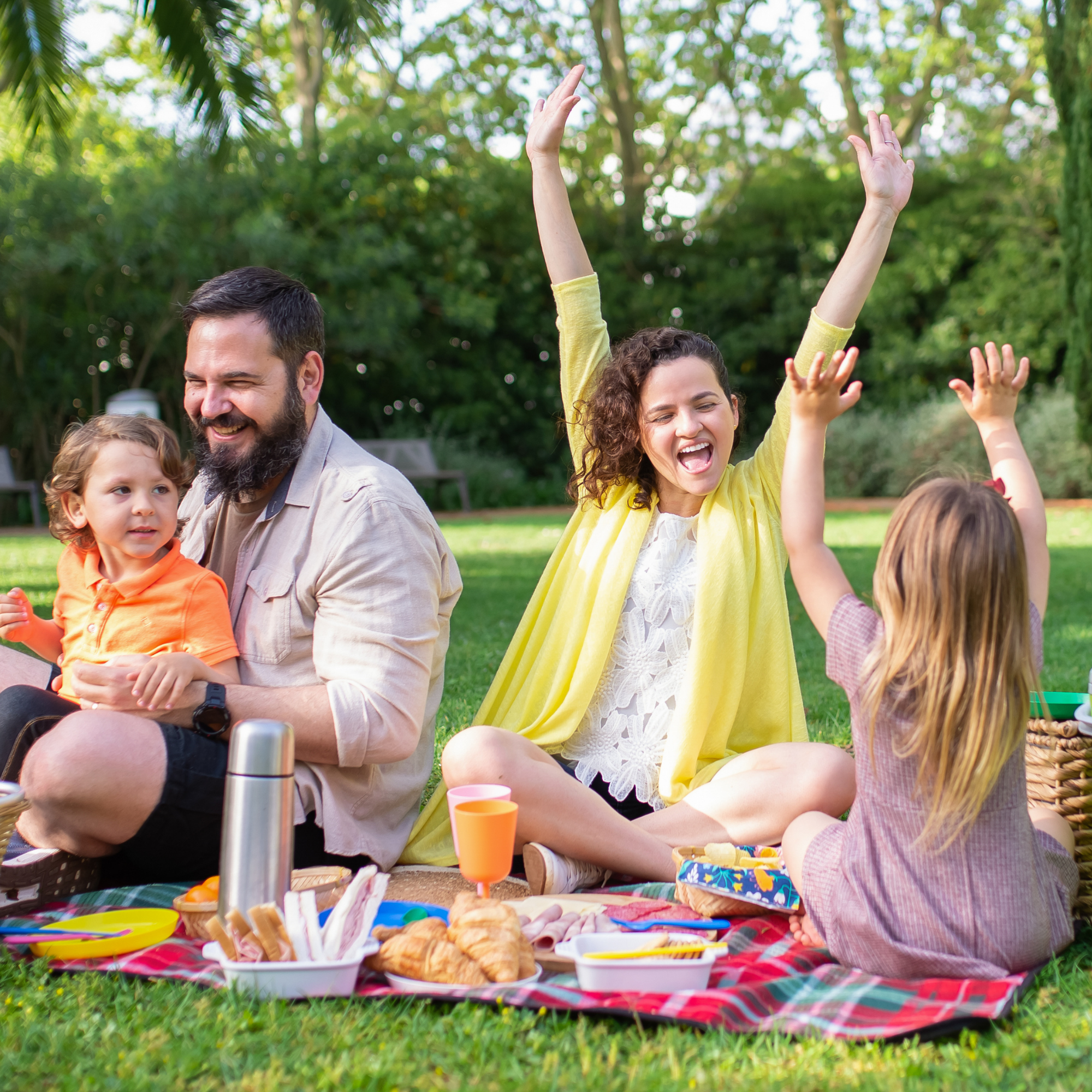 People having a picnic on a checkered blanket in a park, with food and drinks, and children and adults enjoying the outdoor gathering.