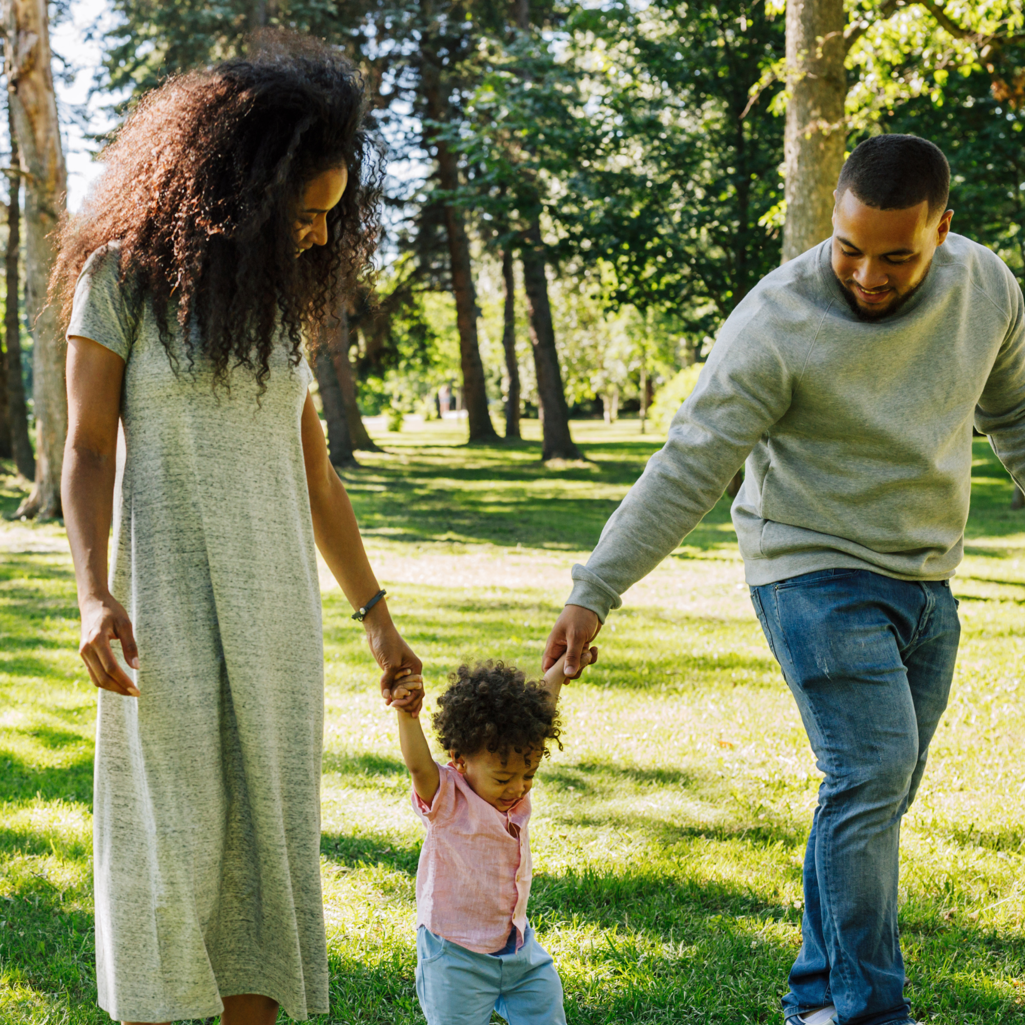 A family of three, including a young girl with curly hair, a woman with long curly hair, and a man with short hair and a beard, are walking hand-in-hand in a park with tall trees and green grass during daytime.