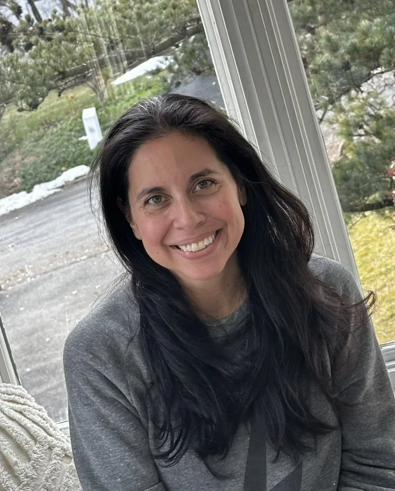 A woman with long dark hair smiling indoors, sitting by a window with a view of trees and a snowy outdoor landscape.