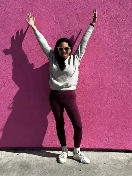 Woman with sunglasses smiling, standing in front of a pink wall, with her arms raised and casting a shadow that makes it look like a dragon or dinosaur is behind her.