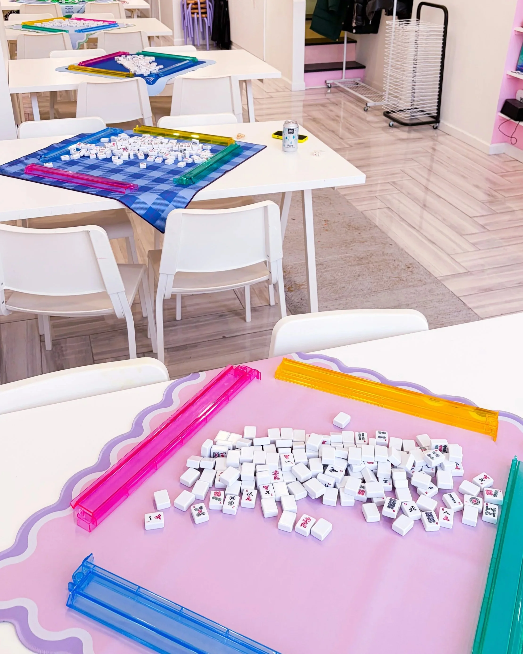 A table in a brightly colored room with Mahjong game pieces scattered across a pink game mat, an orange plastic tile rack, and a clear green tile rack. Other tables in the background display similar games, and the room has wood flooring and chairs.