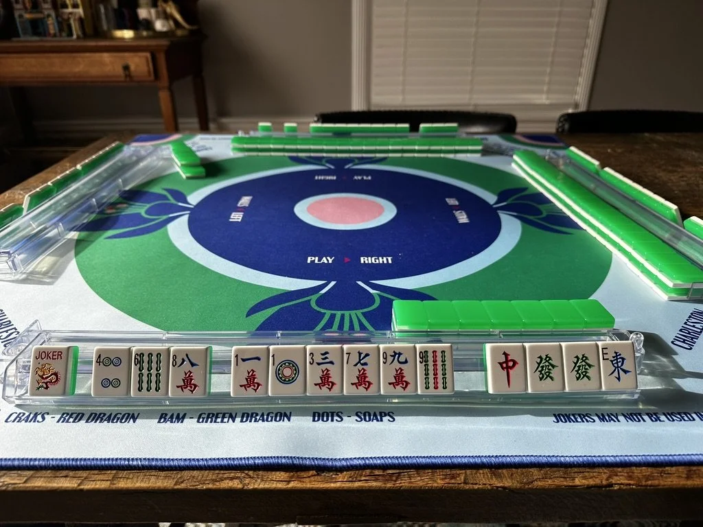 A mahjong game setup on a table with mahjong tiles arranged in a row at the front and green tiles placed around the edge of the mahjong table. The table's surface features a decorative design, and a wooden cabinet with various items is seen in the background.
