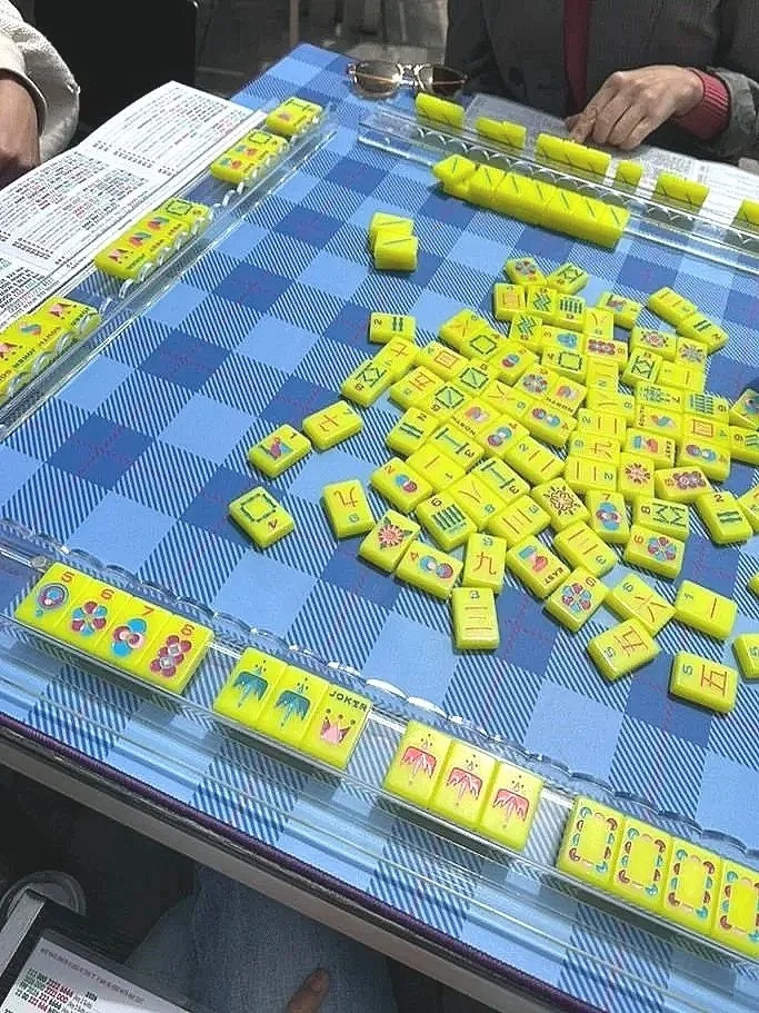 A game of Mahjong with yellow tiles on a blue checkered table. Some tiles are face-up, displaying various symbols and numbers, while others are face-down or stacked. There are players' hands and papers around the table.
