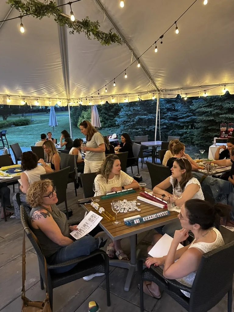 People playing a board game at a table under a decorated outdoor event tent with string lights, while others are seated at tables in the background, and trees are visible outside.