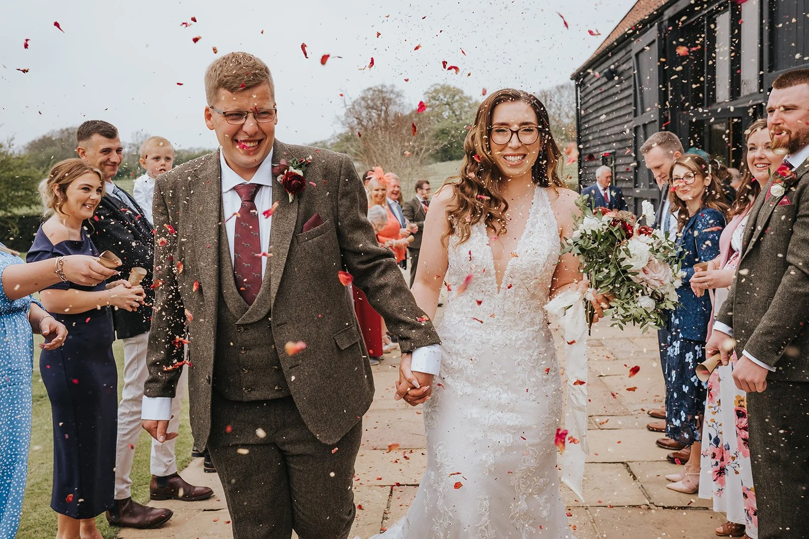A newlywed couple holds hands and walks down the aisle, smiling, surrounded by guests throwing flower petals at an outdoor wedding ceremony.