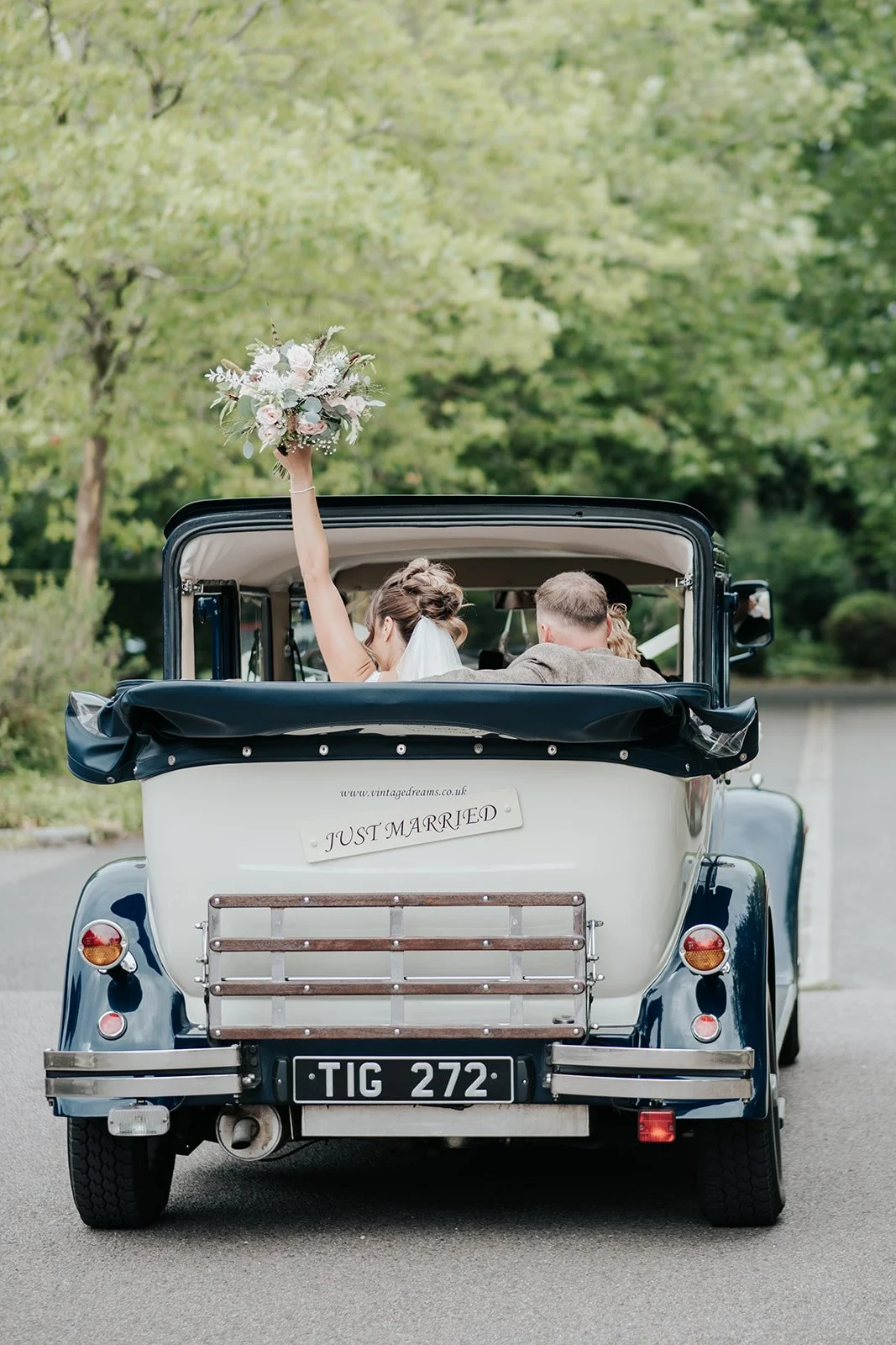 A vintage car with a 'JUST MARRIED' sign on the back, carrying a newlywed couple, with the bride holding a bouquet and waving, driving on a tree-lined road.