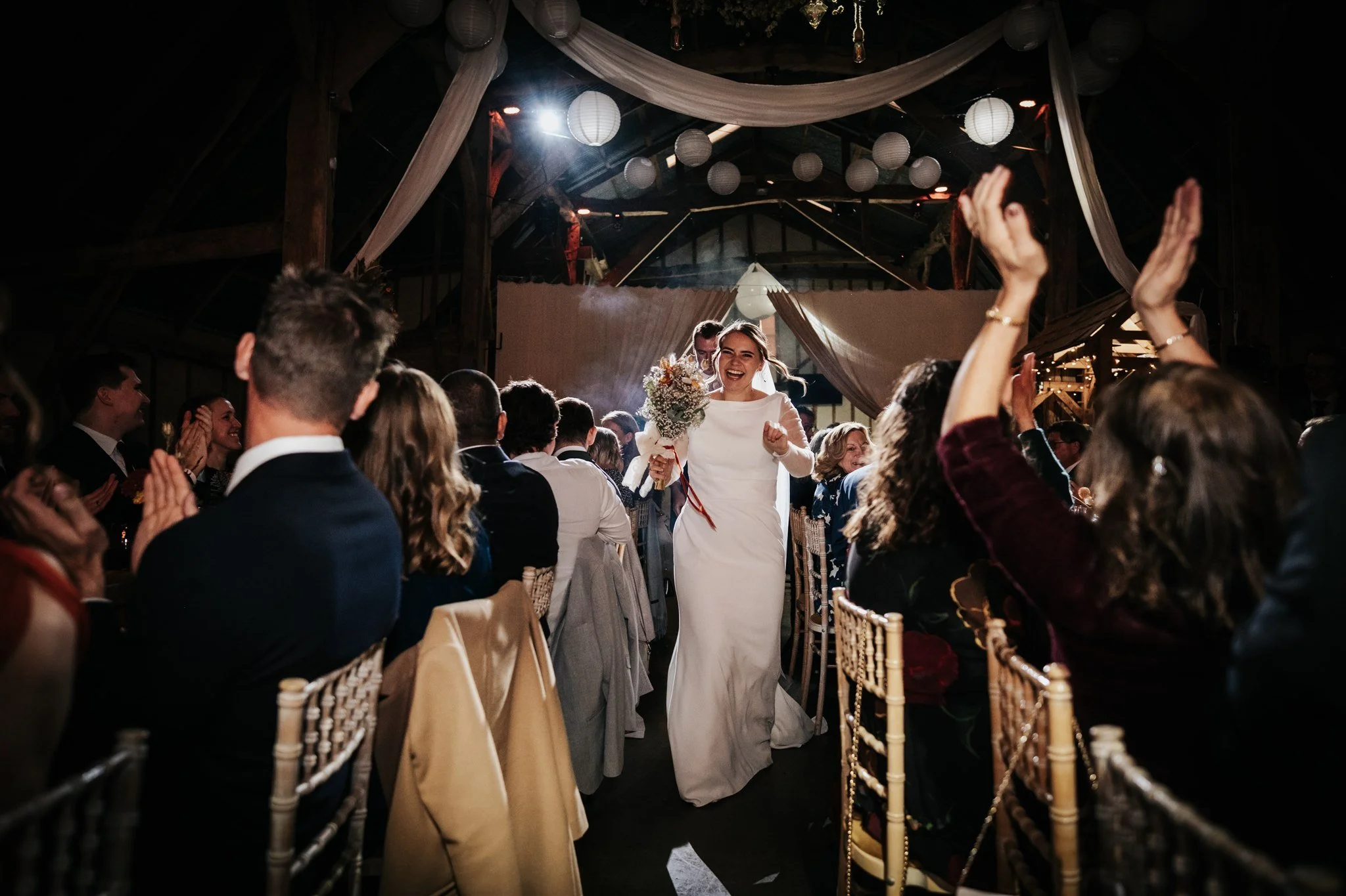 A bride holding a bouquet of flowers smiling and walking down the aisle at her wedding reception, surrounded by guests seated on both sides, in a decorated venue with hanging lanterns and draped fabric.