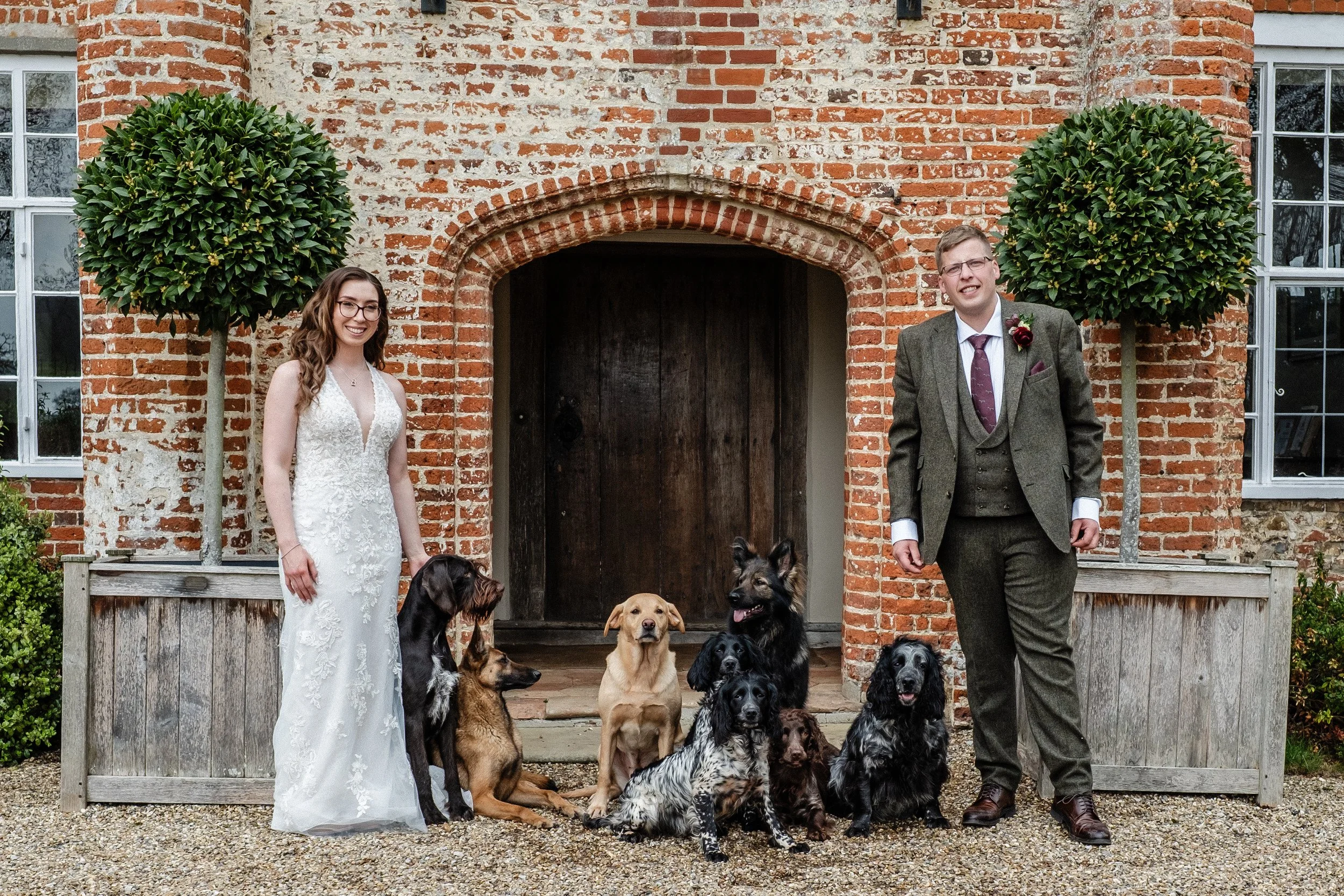 A bride and groom standing in front of a brick building with two windows, surrounded by onlookers and dogs, at a wedding at Bruisyard Hall, Suffolk.