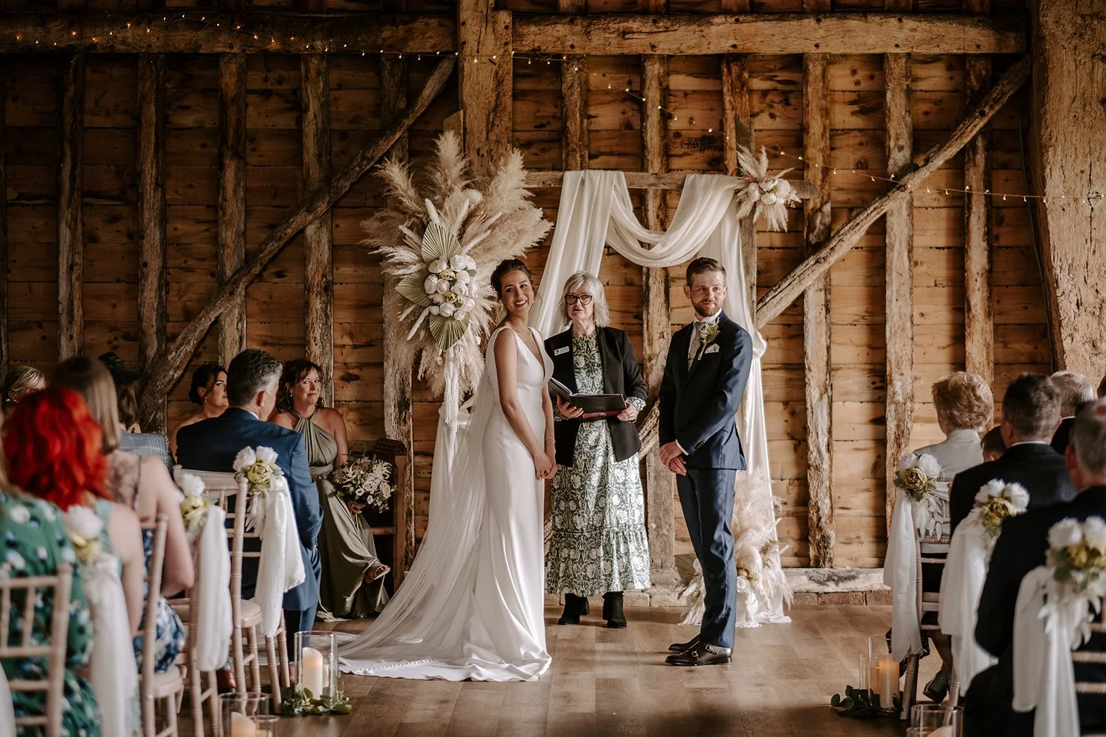 A wedding ceremony at Suffolk Barn, Hawstead, Suffolk with a bride and groom facing a registrar , standing in front of a rustic wooden backdrop decorated with large floral arrangements. 