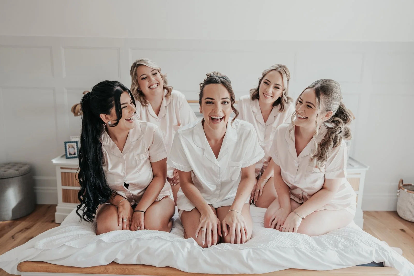 A bride with her bridesmaids in pyjamas on a bed, smiling and laughing at each other in a bright, modern bedroom.