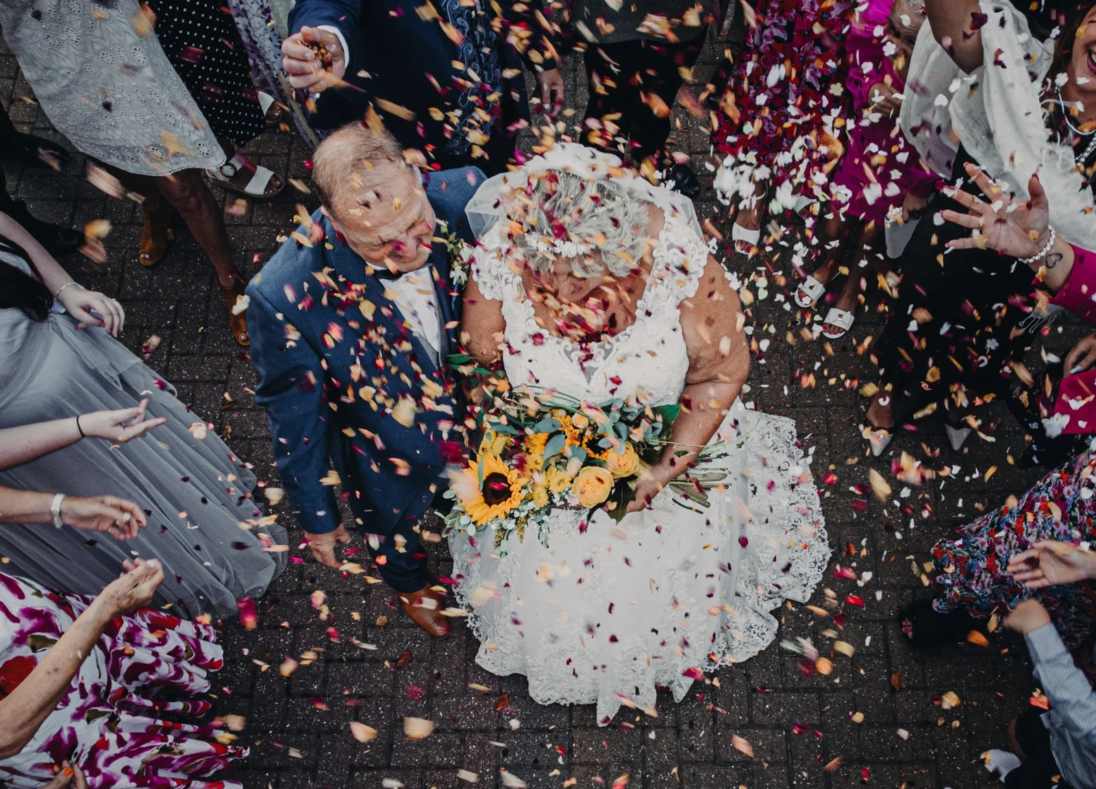 Bride in a white lace dress holding a bouquet of yellow and orange flowers, walking among guests throwing flower petals, at a wedding celebration.