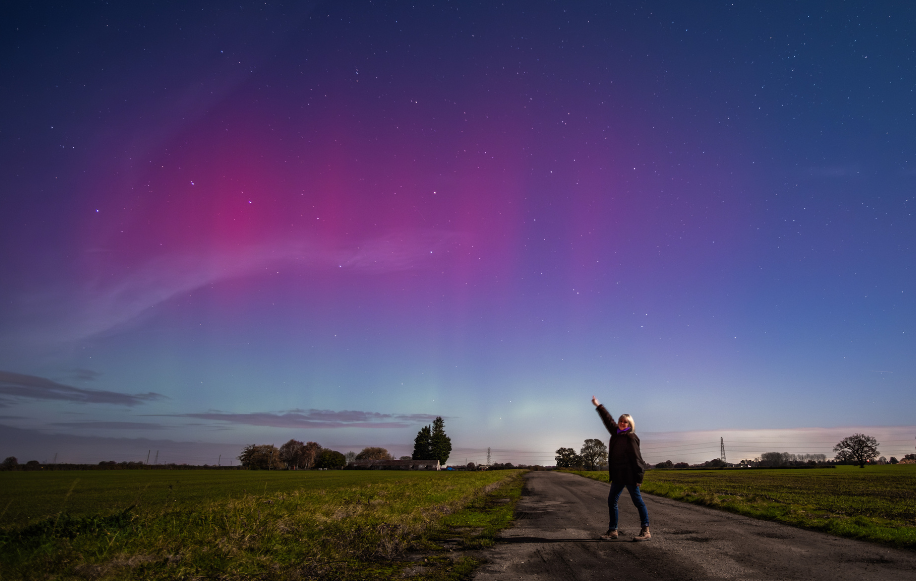 Person standing on a rural road at night, pointing towards the colorful aurora borealis and starry sky above.